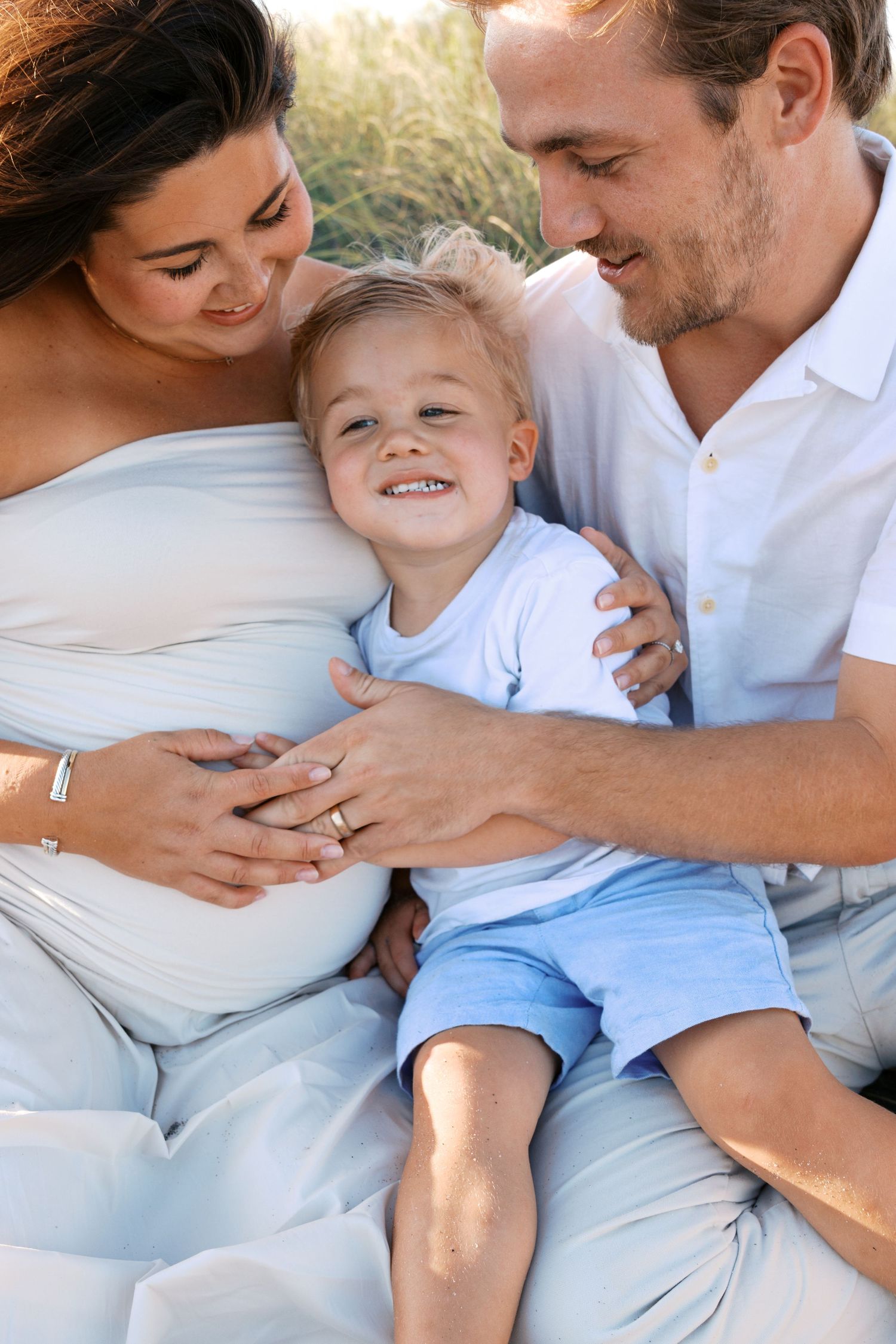 Smiling family with pregnant mother, father, and young son share a joyful moment together on a beach.