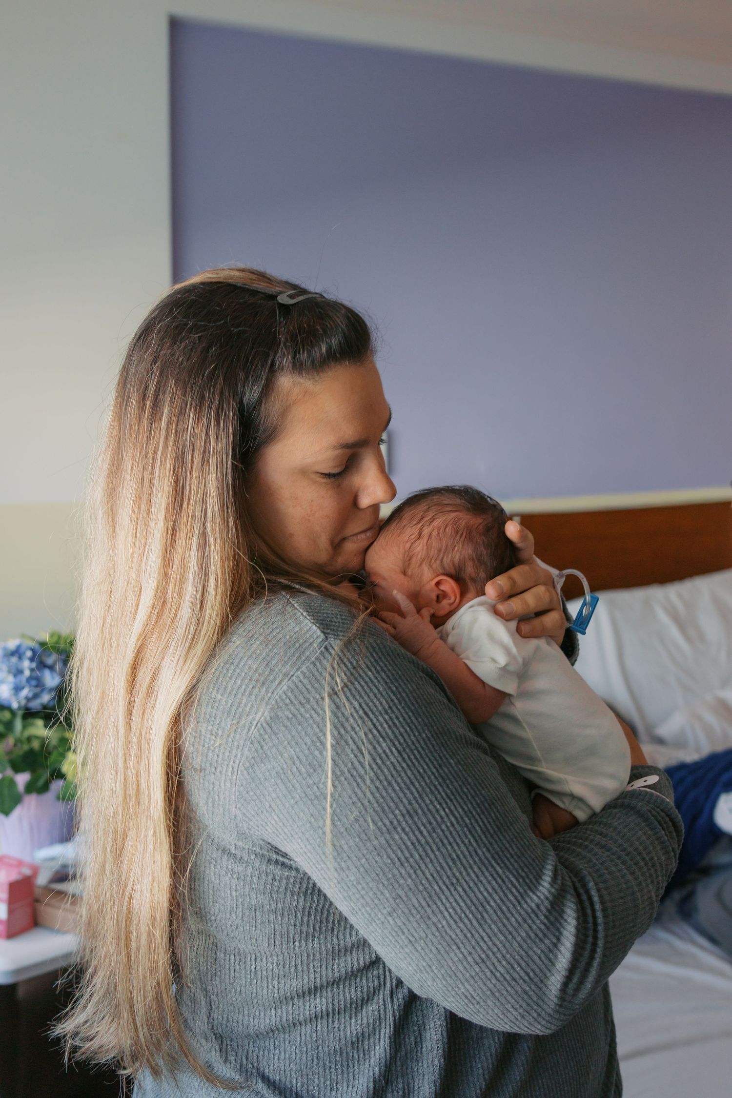A mother tenderly holds and kisses her newborn baby in a hospital room after giving birth.