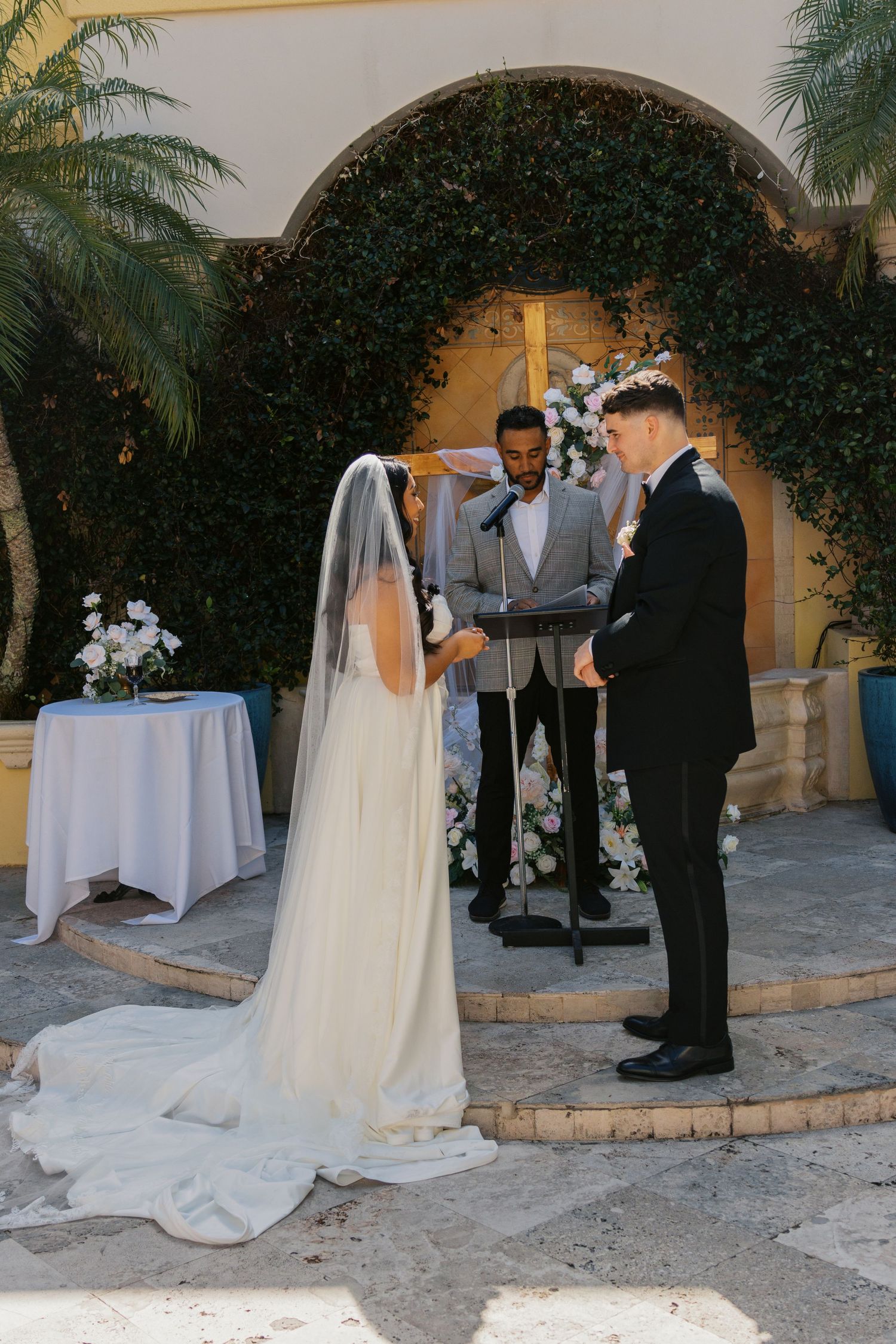 Bride and groom exchange vows at outdoor wedding ceremony beneath lush ivy arch with white floral arrangements.
