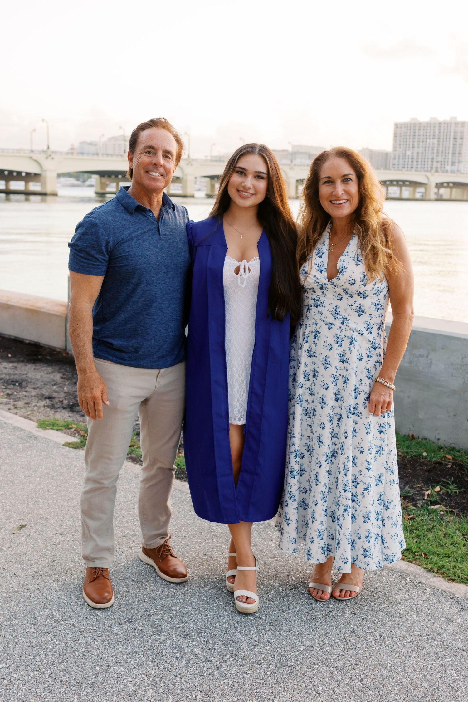 Family of three posing outdoors near waterfront, graduate in blue gown flanked by proud parents on sunny day.