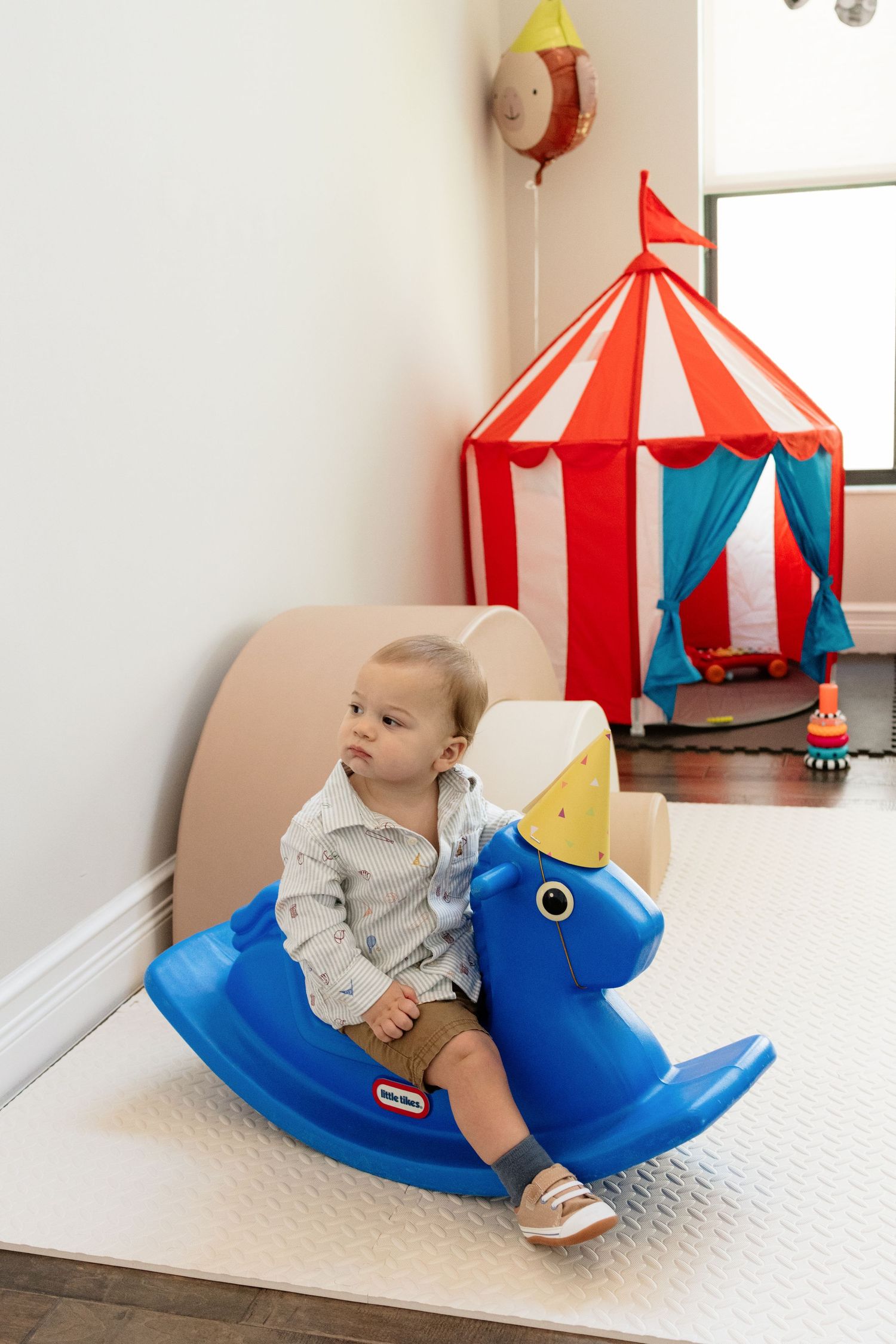 Baby boy sitting on a blue Little Tikes rocking horse toy near a red and white striped circus play tent.