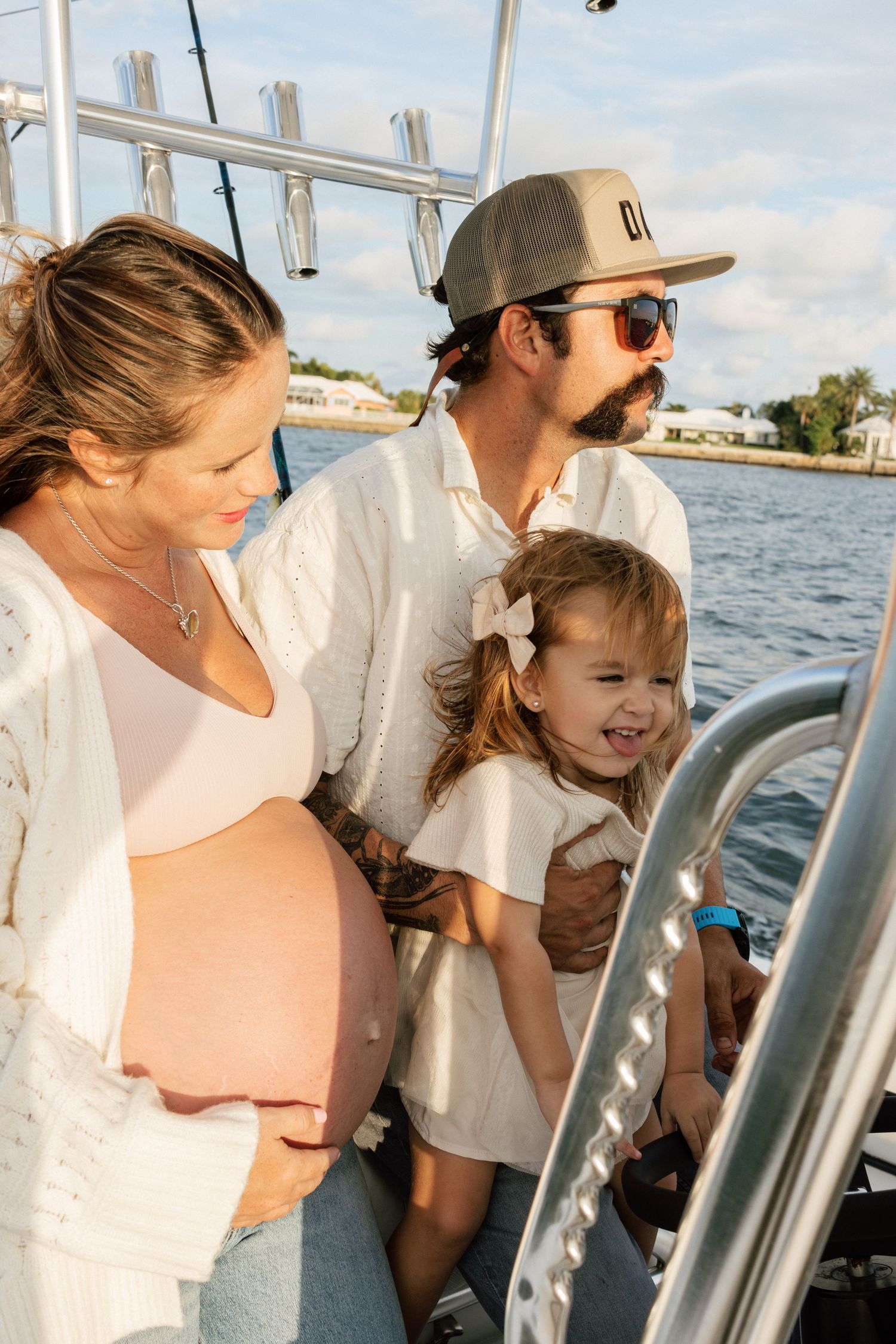 Pregnant woman, bearded man in cap and sunglasses, and smiling toddler girl enjoy time together on a boat near the water.