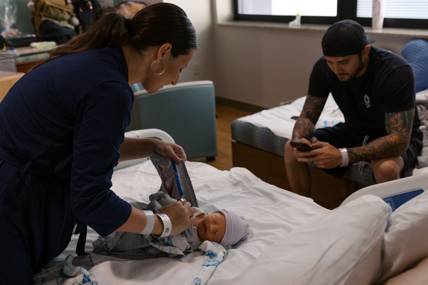 Parents caring for newborn baby in hospital room, mother changing diaper while father uses smartphone.