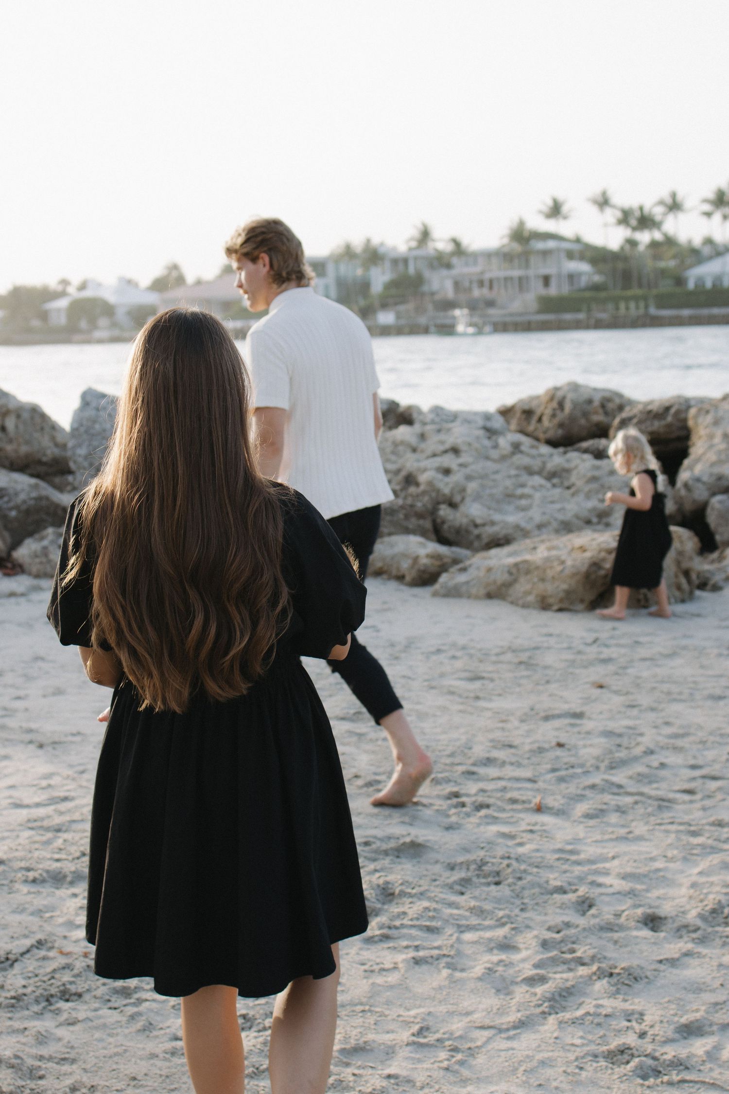 People walking along a sandy beach with coastal homes visible in the background at sunset.