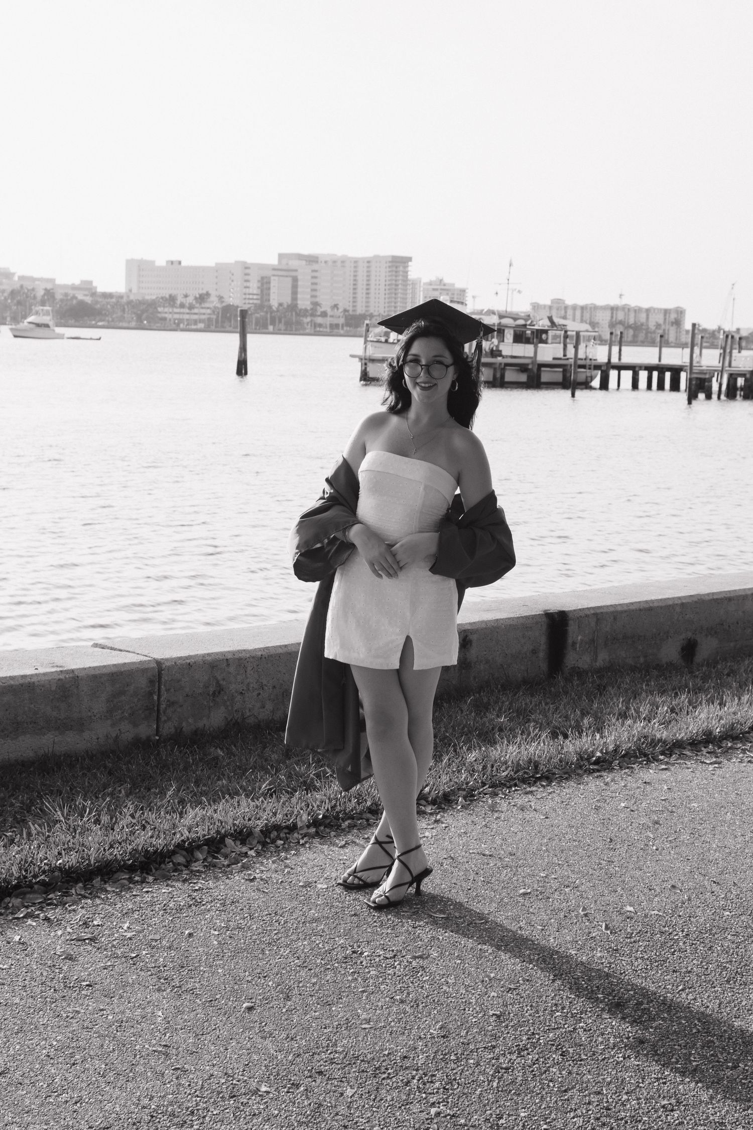 Graduate in white dress and cap poses by waterfront dock.