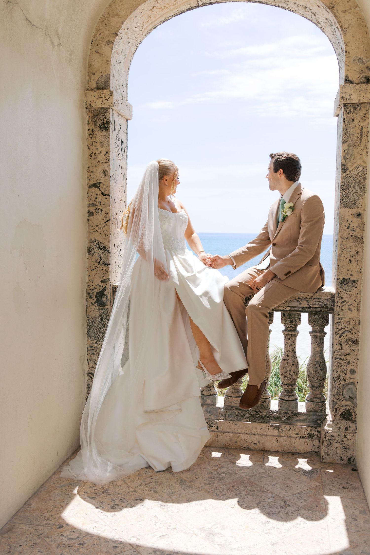 Bride and groom hold hands in a stone archway overlooking the ocean on their wedding day.