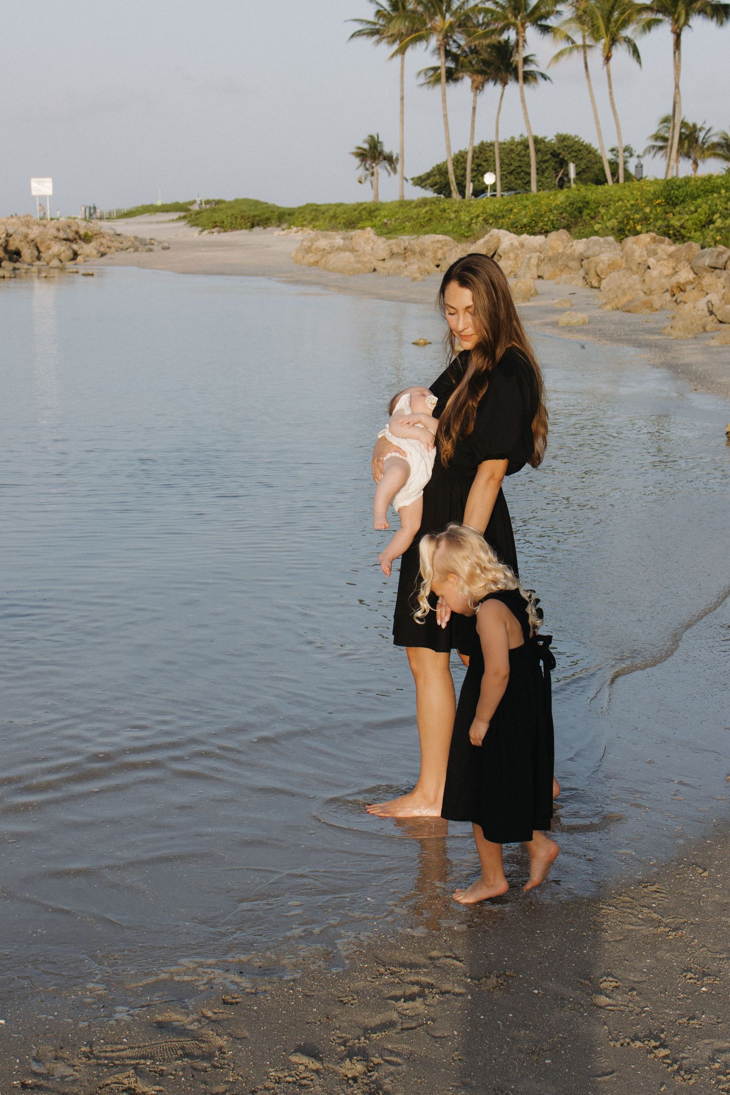 A person stands in shallow water at the beach holding a small child with palm trees in background.