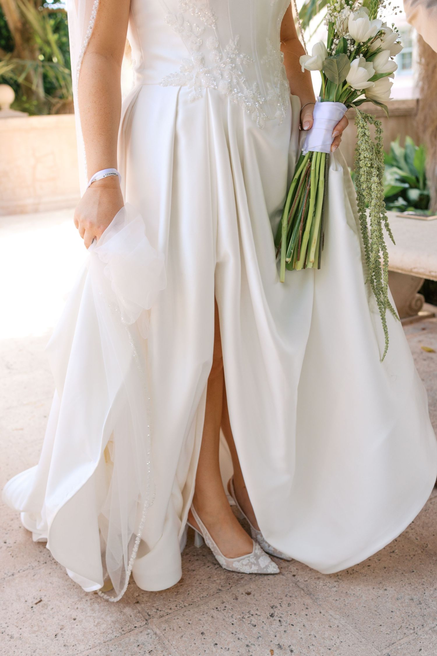 Bride in white satin wedding gown with slit holding elegant white floral bouquet with greenery, wearing silver heels.