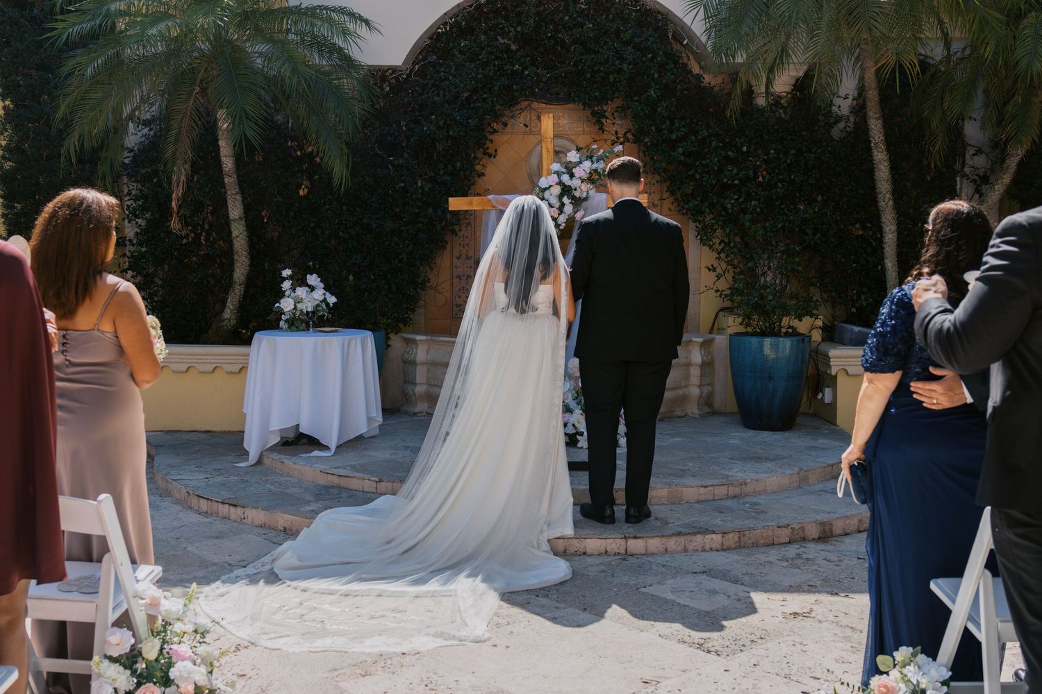 Bride and groom standing at outdoor wedding altar with guests seated, lush greenery and floral arrangements in background.