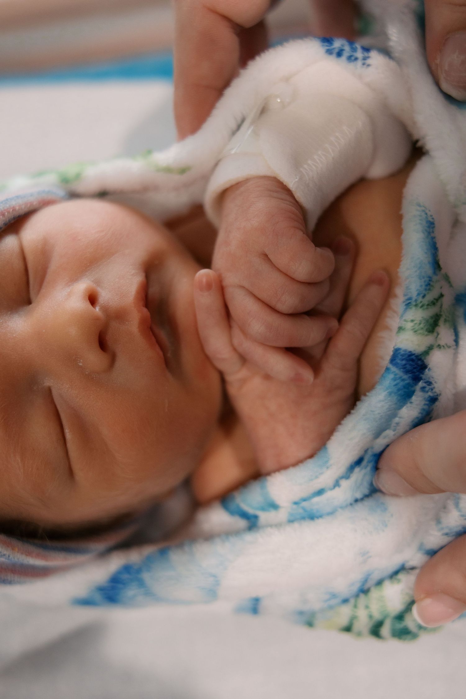 Newborn baby sleeping peacefully wrapped in a blue and white blanket, holding an adult finger.