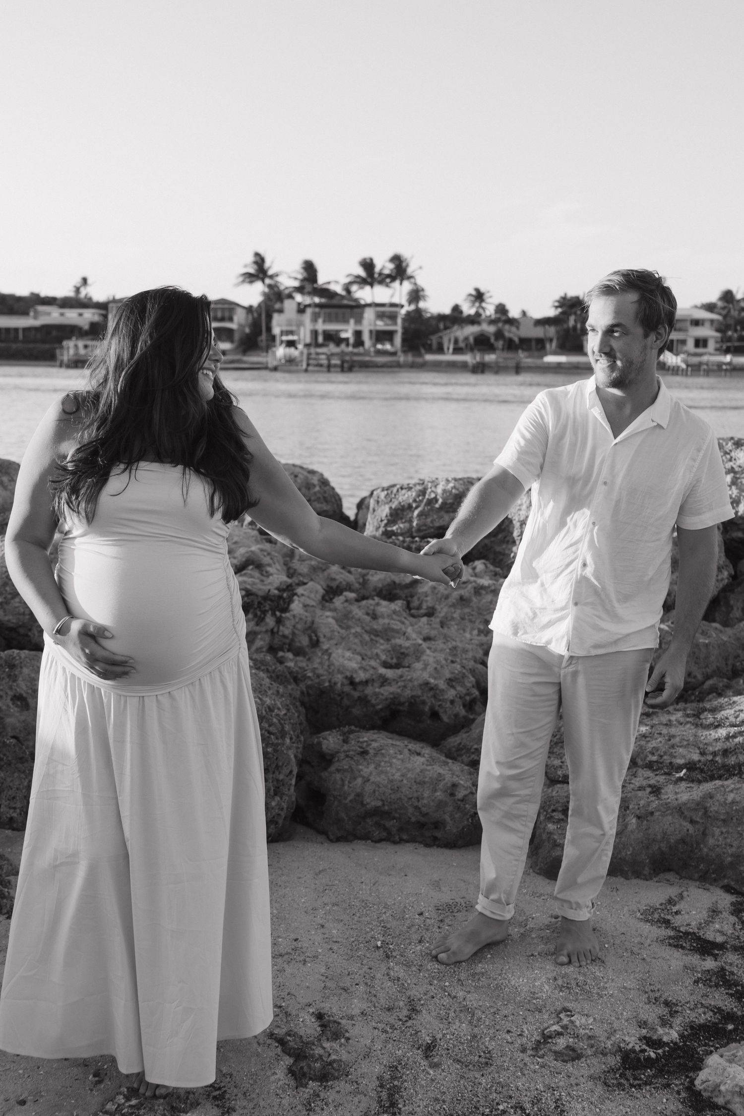 Black and white photo of pregnant couple holding hands on rocky beach with waterfront homes behind them.