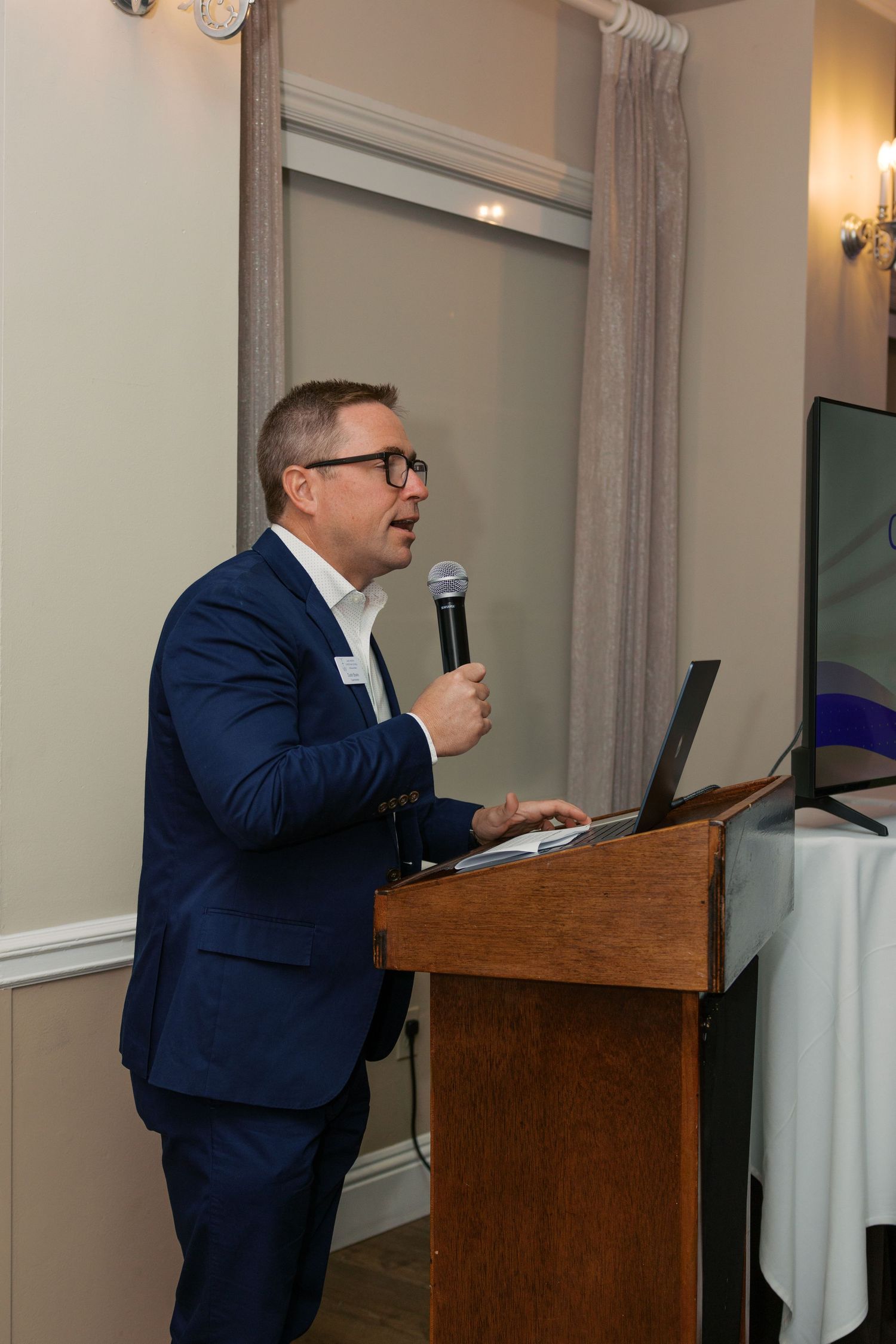 Man in blue suit speaking at podium with microphone and laptop during professional conference presentation.