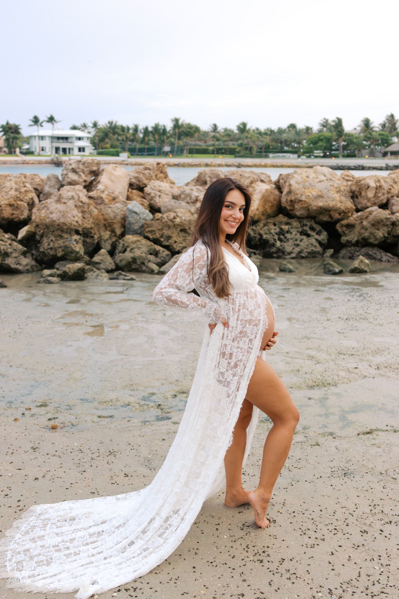 Pregnant woman in white lace maternity gown posing on a rocky beach shore with palm trees in the background.
