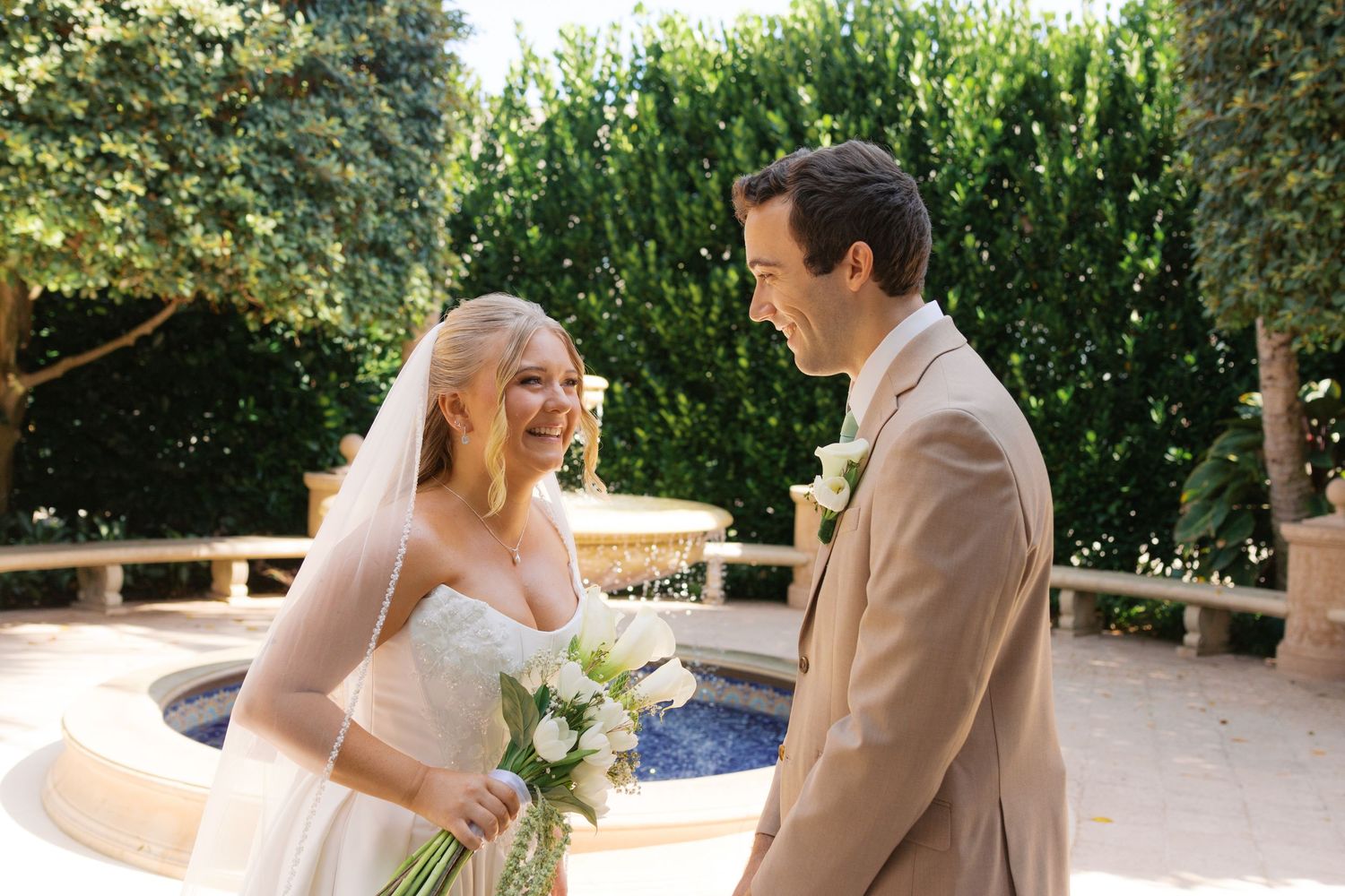 Bride and groom share a joyful moment outdoors, with bride holding white bouquet near a fountain.