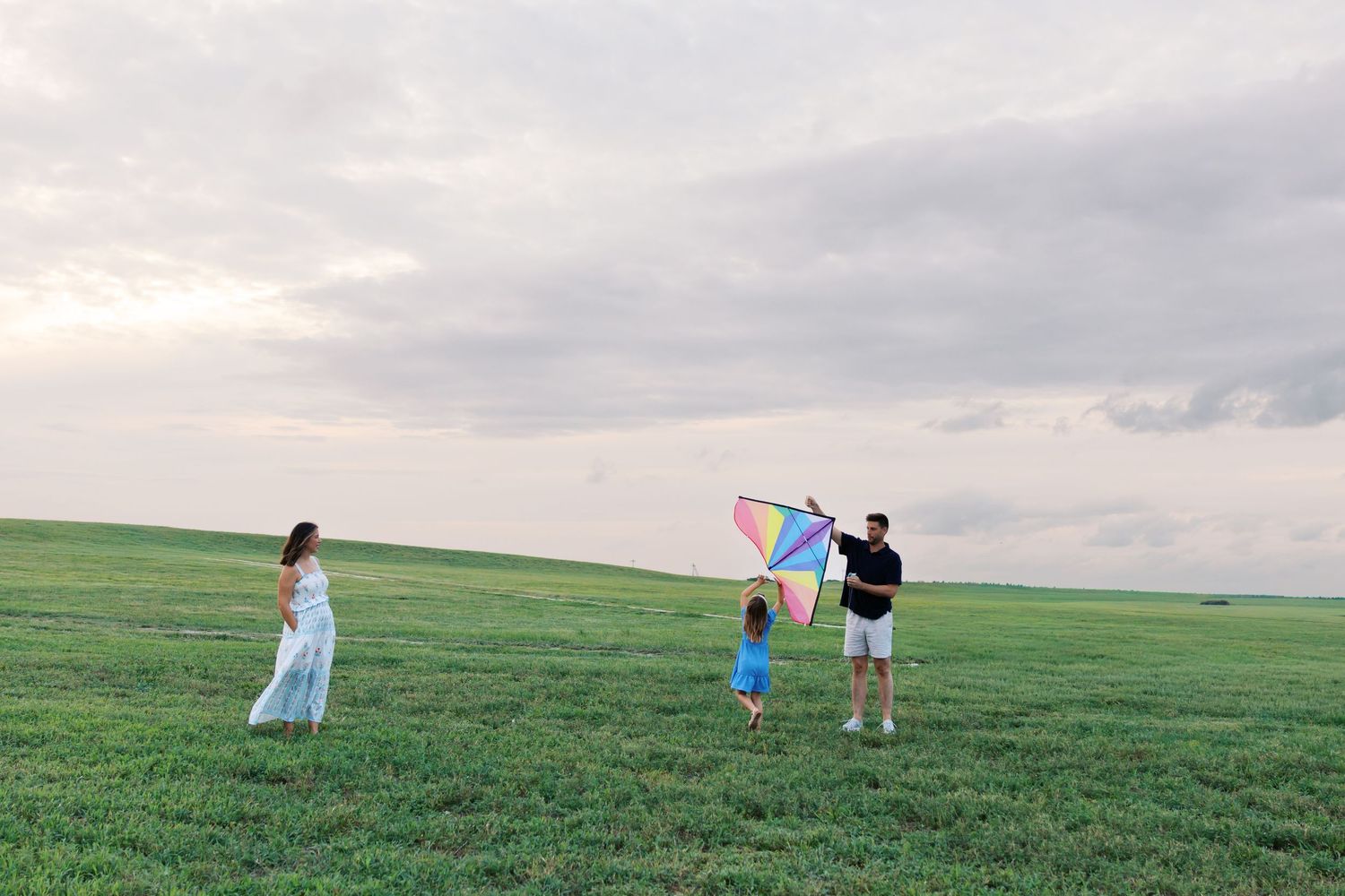 Family flying a colorful kite together in a vast green field under a cloudy sky.