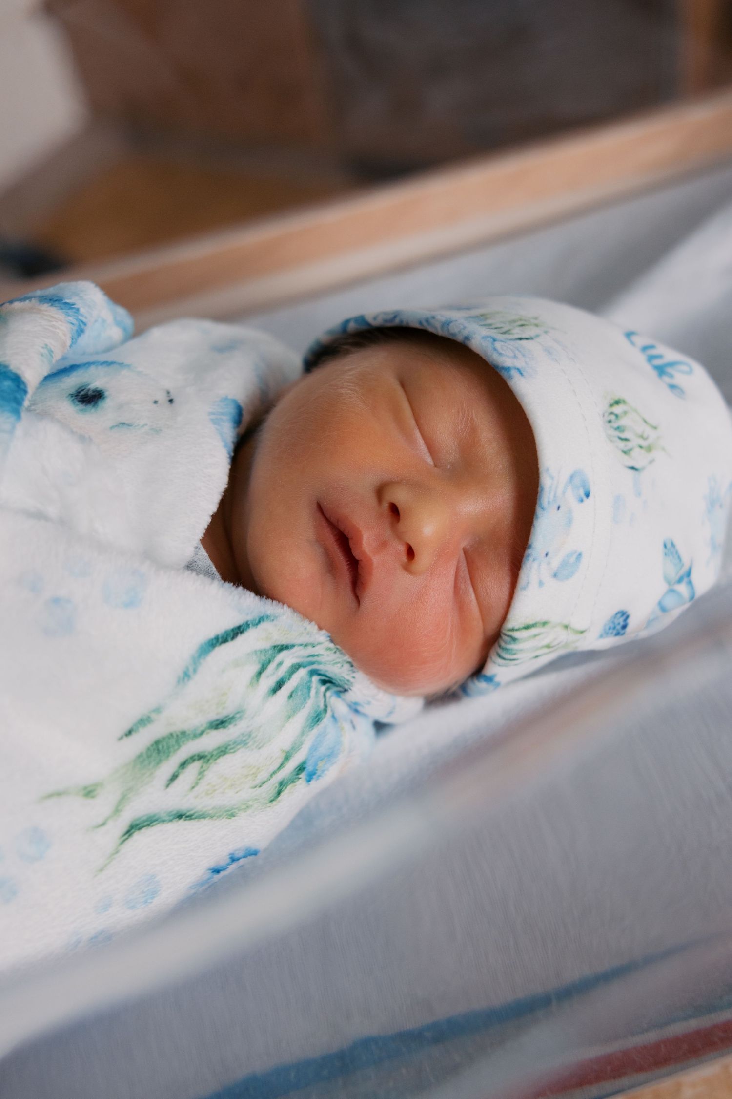 Newborn baby sleeping peacefully swaddled in white blanket with blue pattern, wearing matching hat in hospital bassinet.