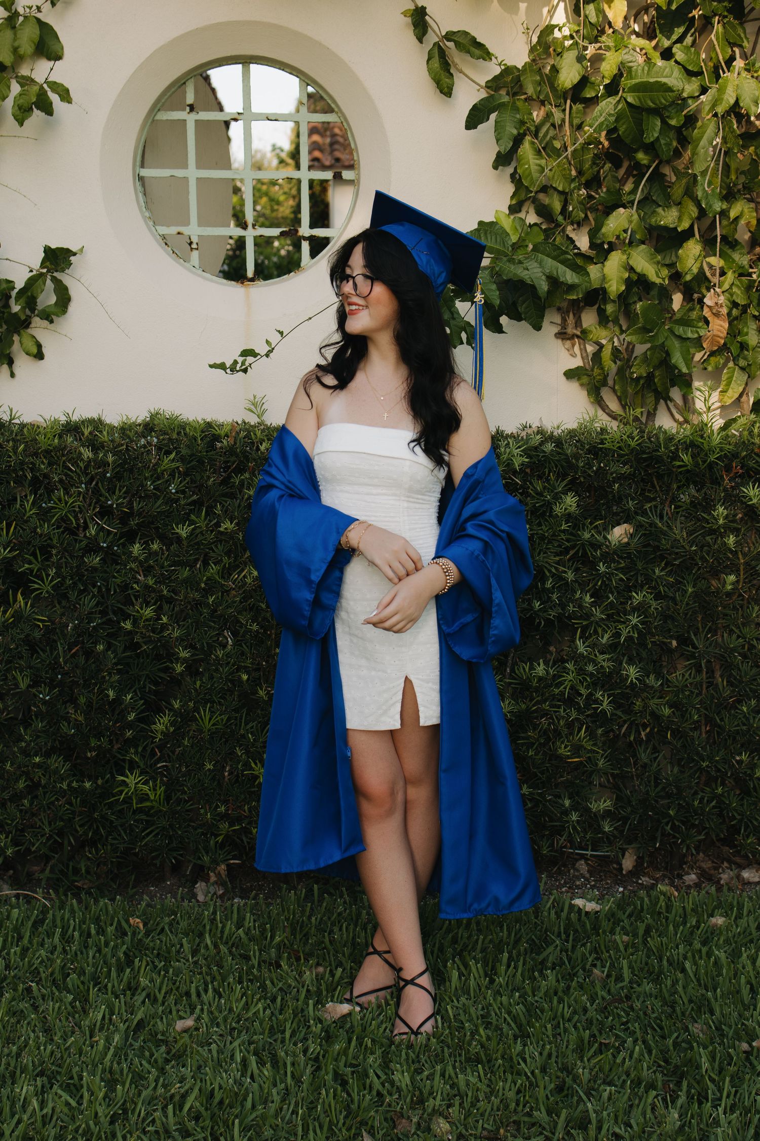 Graduate in blue cap and gown poses against ivy-covered wall with arched windows.