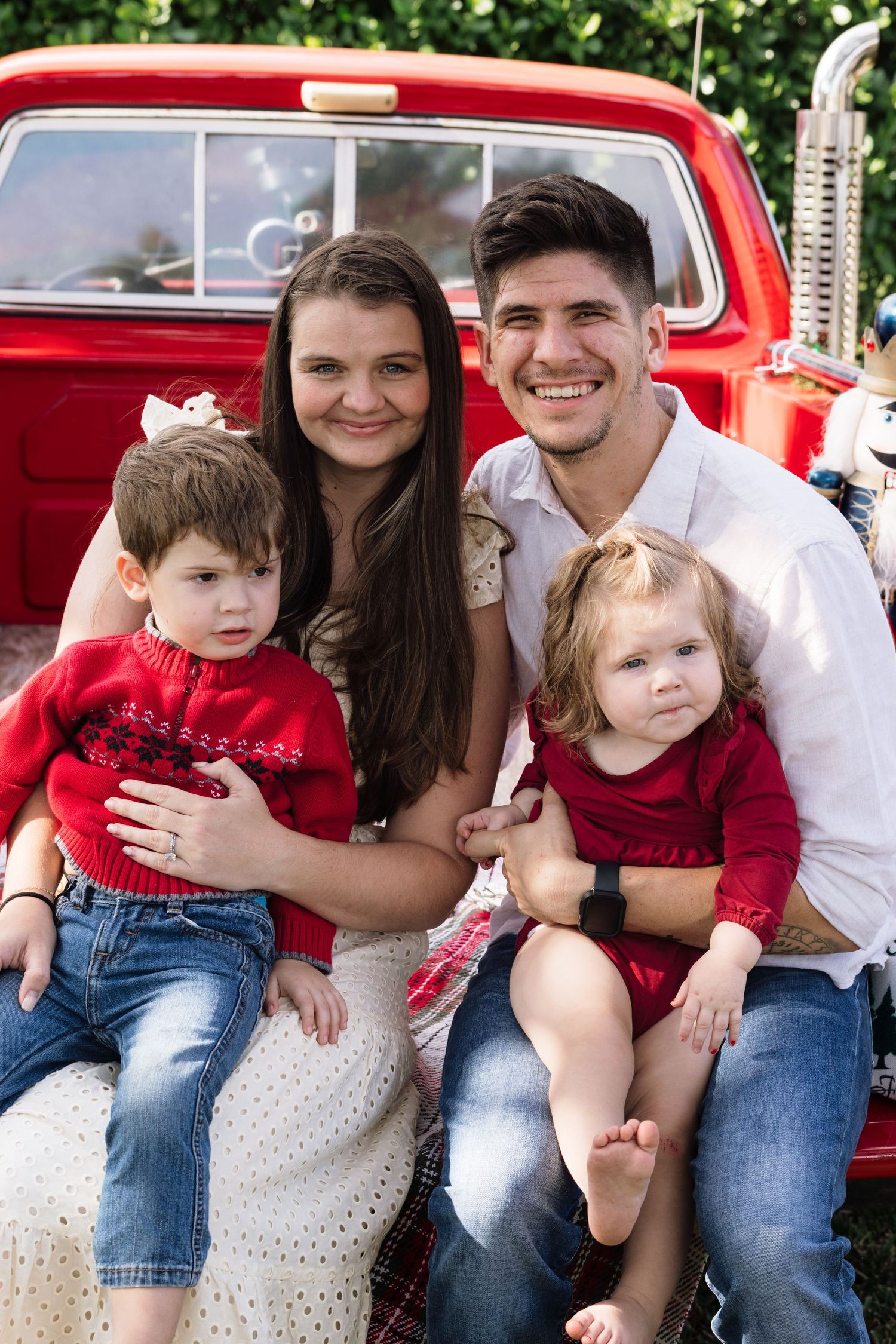 A smiling young family with two toddlers in red outfits poses on a vintage red pickup truck tailgate outdoors.