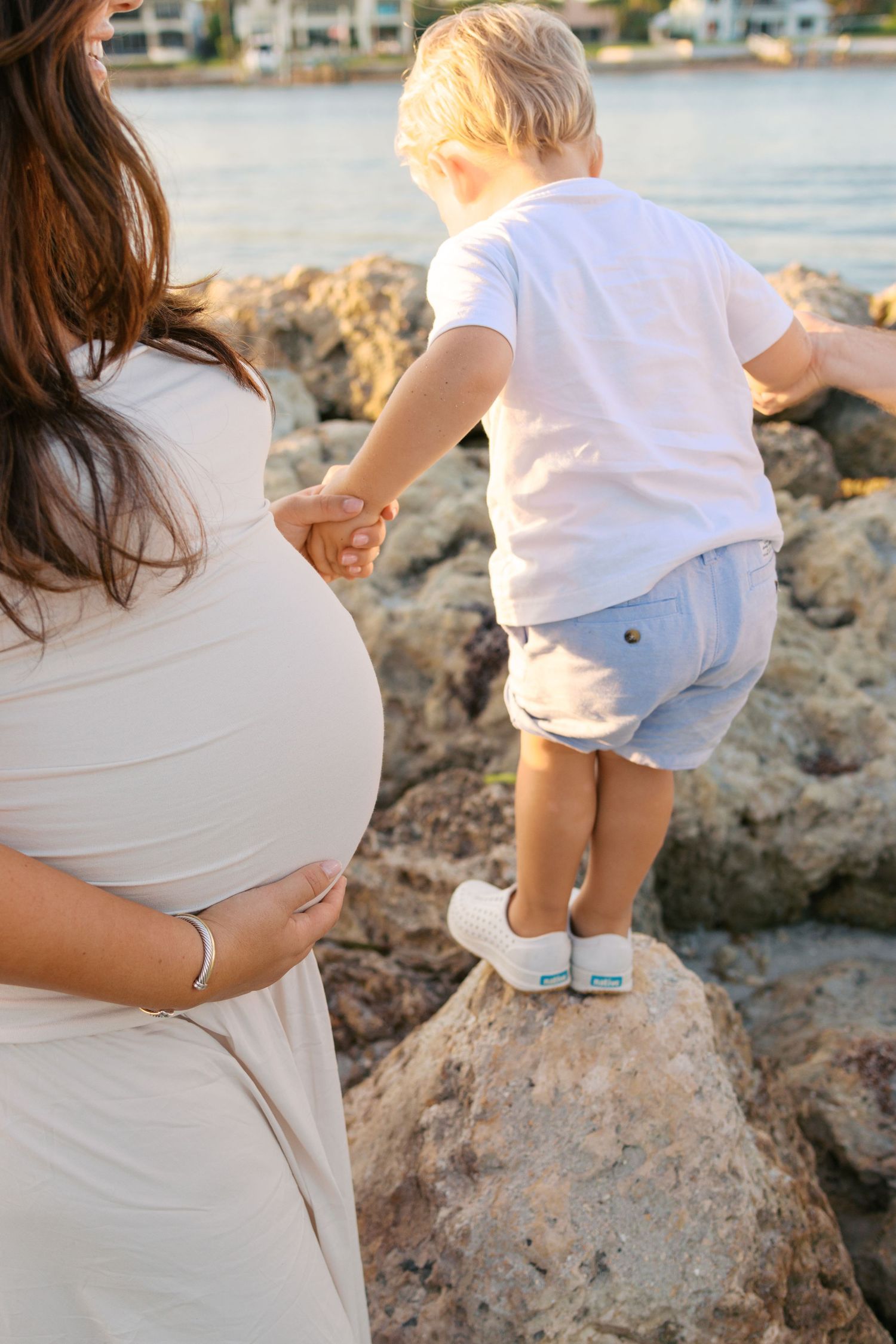 Pregnant woman in beige dress holds toddler boy's hand as he stands on rocks by the water.