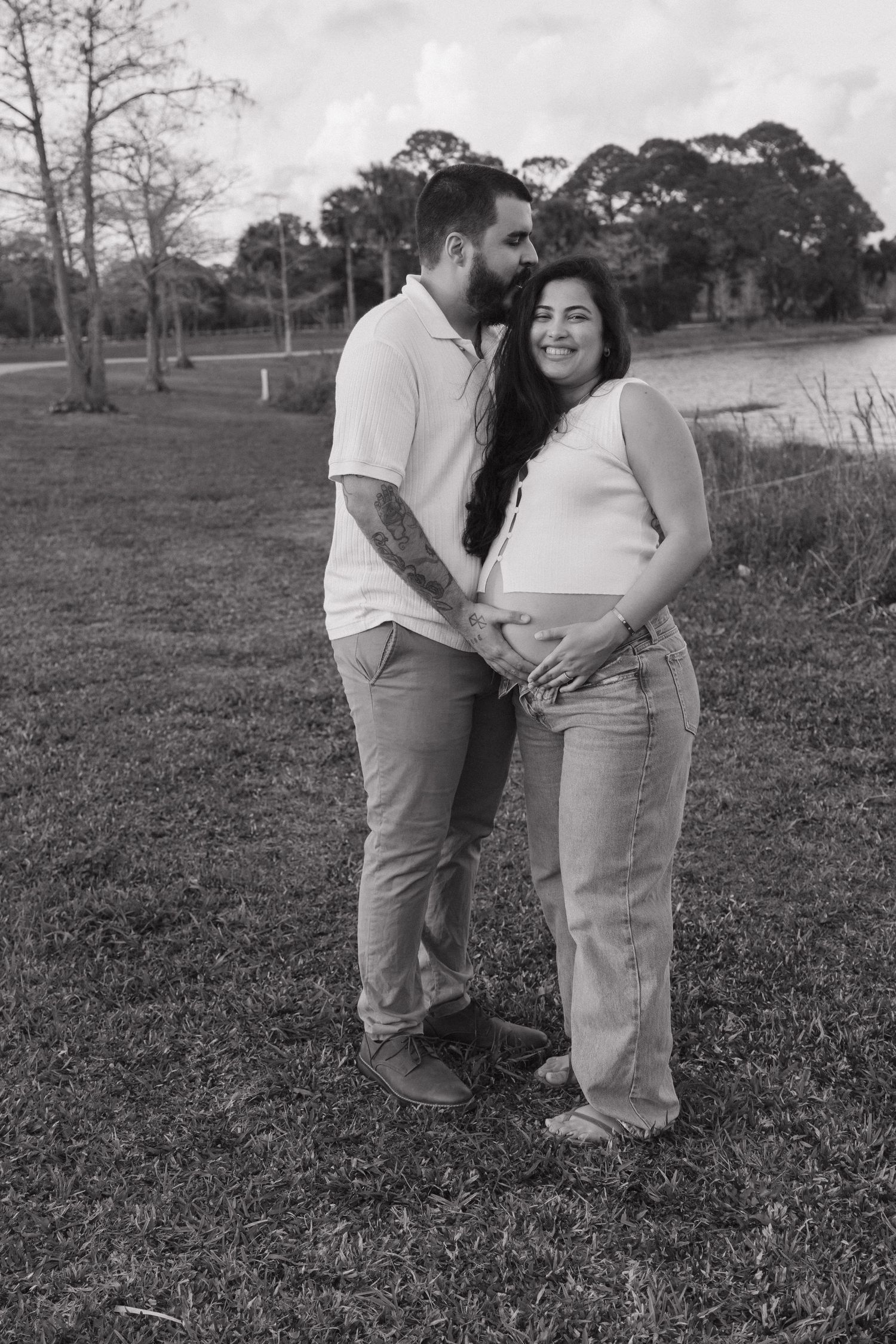 Black and white photo of a couple outdoors, man kissing pregnant woman's cheek while both cradle her baby bump.