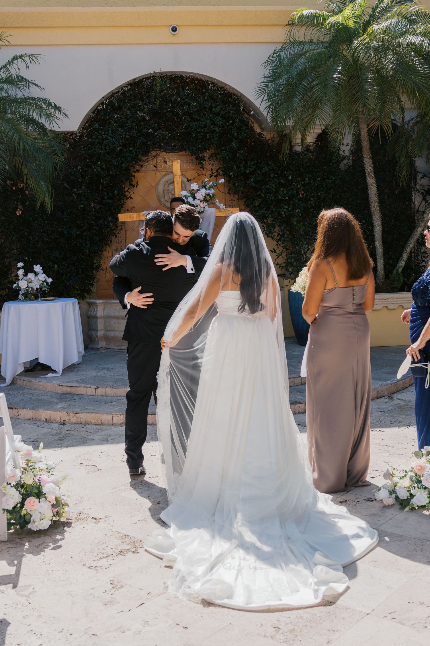 Bride in white gown and veil embraces groom at outdoor wedding ceremony with lush greenery backdrop.