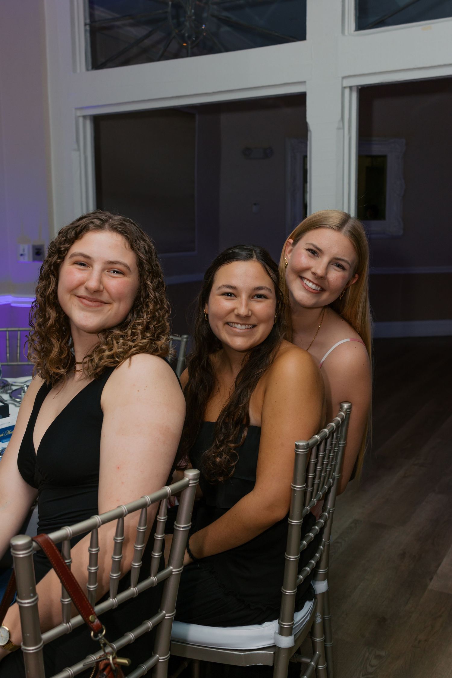 Three young women smiling and posing together at a formal event, seated on silver chiavari chairs.