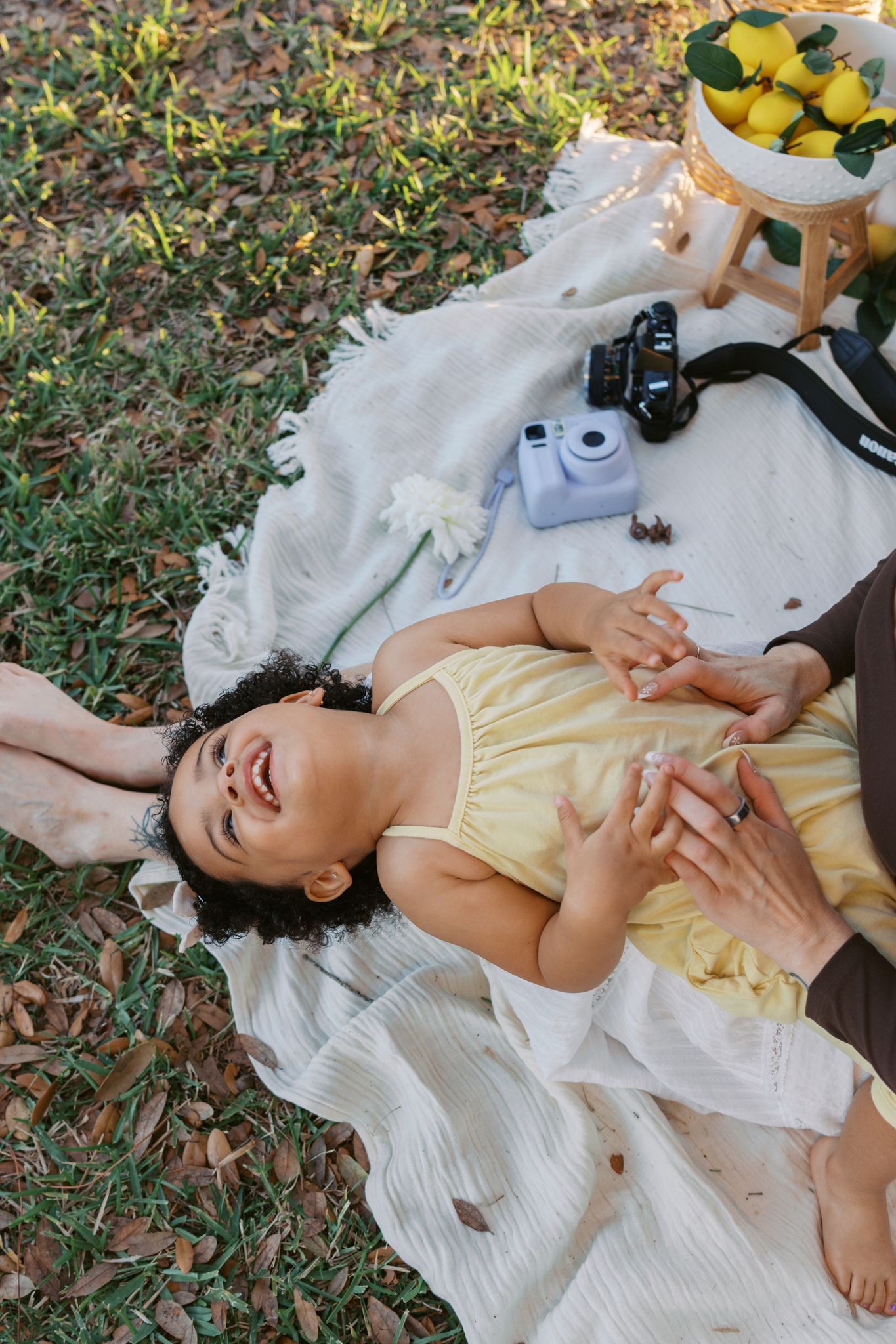 A laughing toddler in a yellow dress lies on a blanket outdoors while an adult plays with them near a camera and lemons.