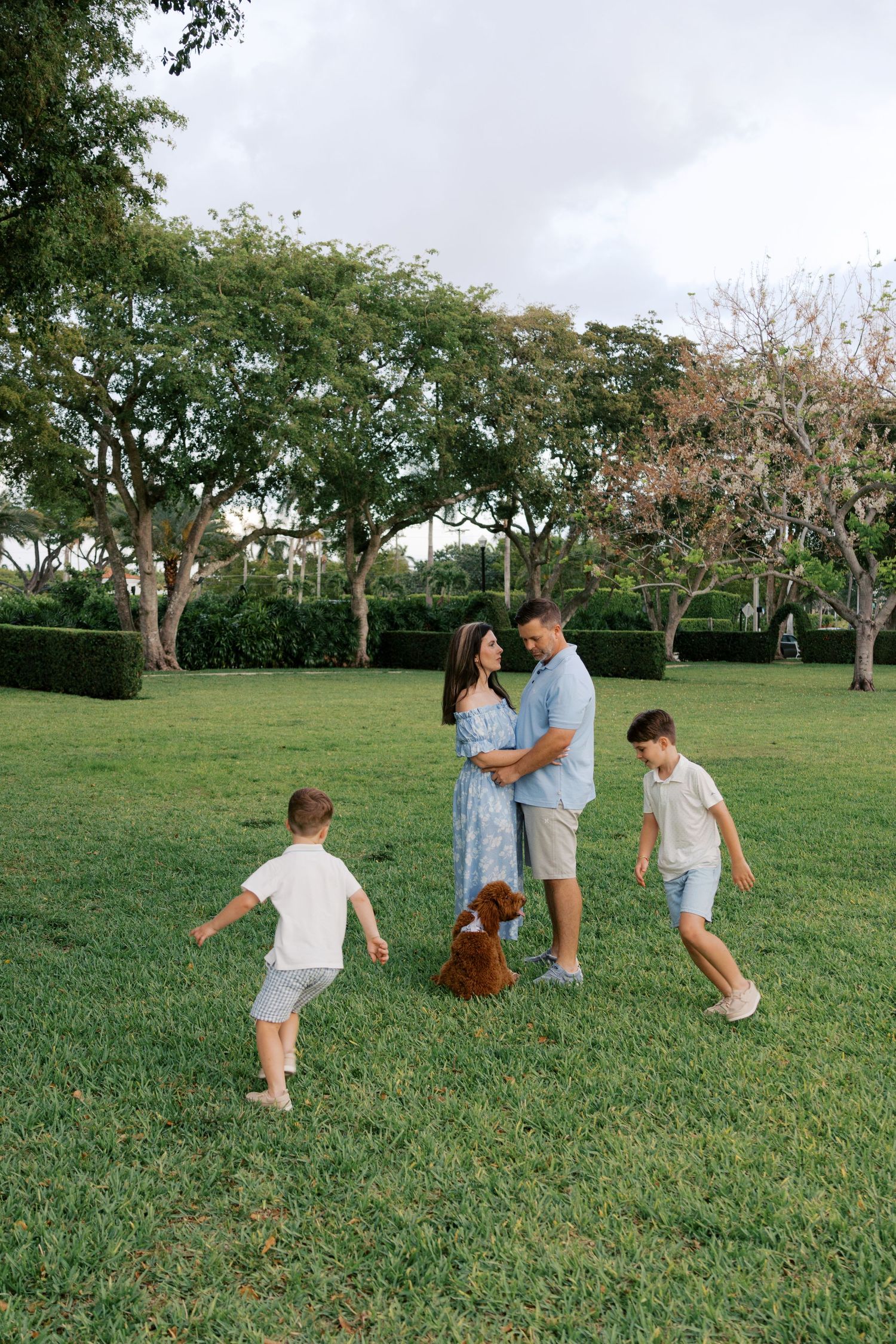 Family with two boys and a dog enjoying a sunny day on green grass in a park with lush trees.
