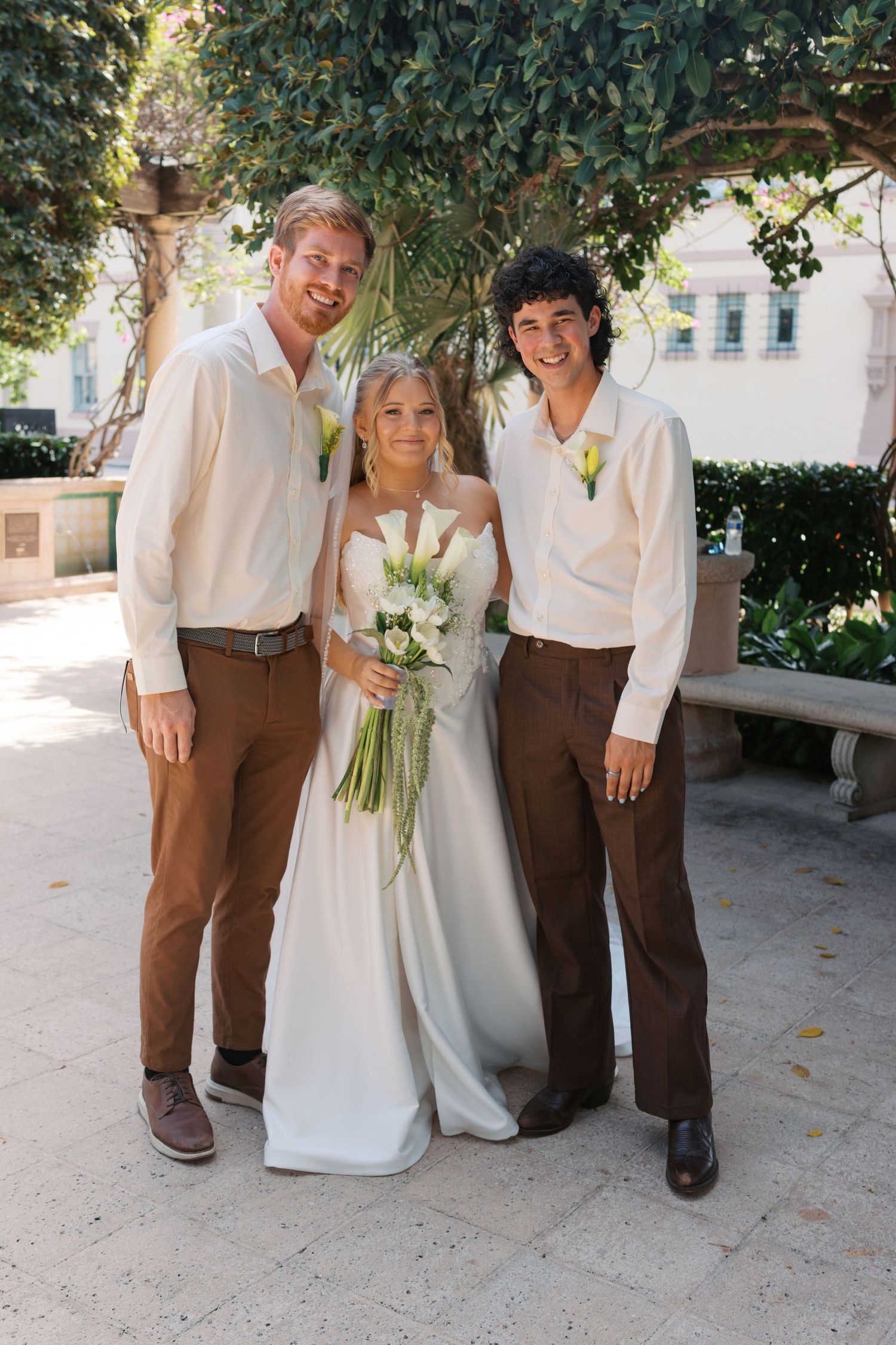 Bride in white gown holding bouquet poses with two groomsmen in brown pants and cream shirts outdoors.