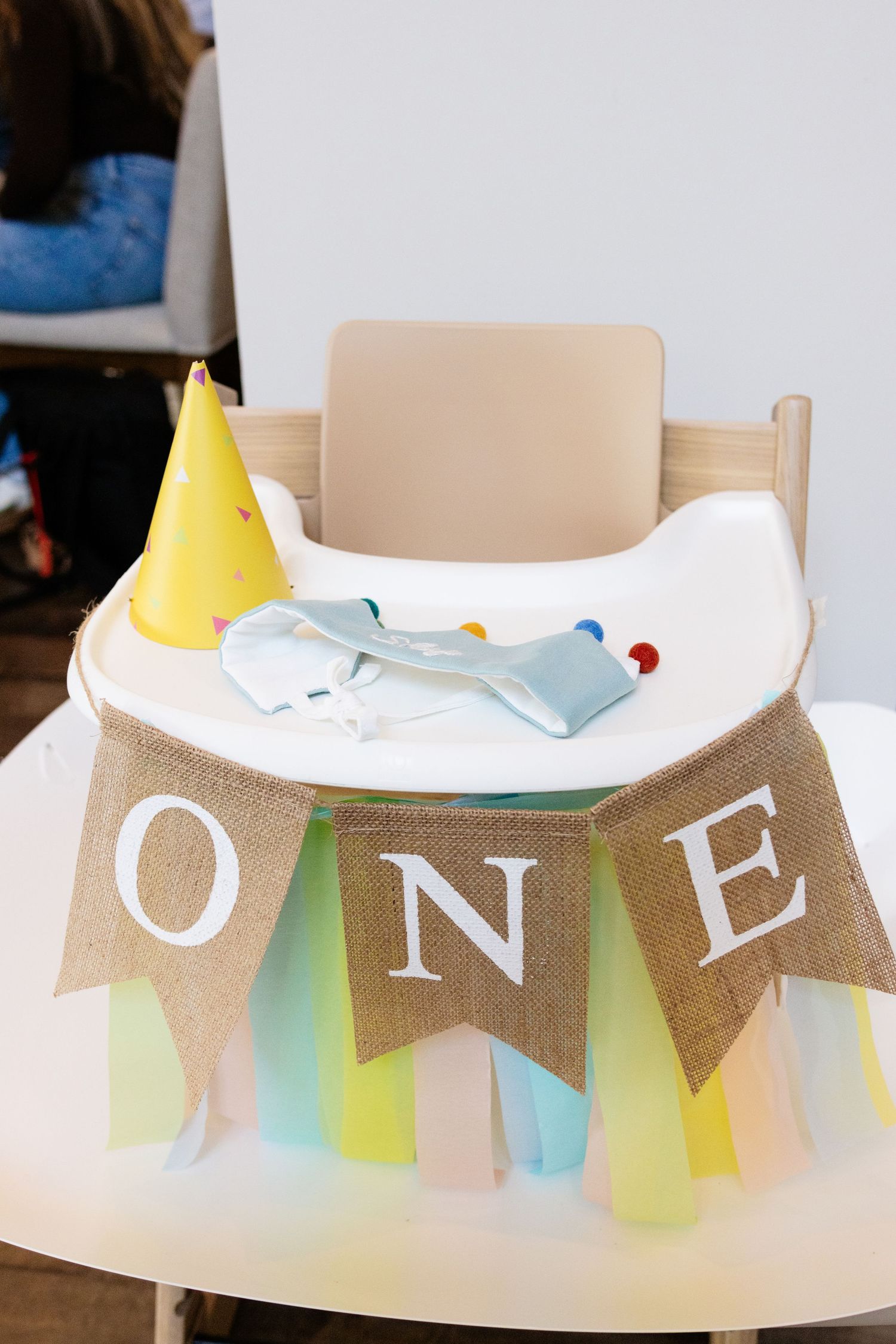 A wooden high chair decorated with a burlap ONE banner and pastel tulle skirt, with a yellow party hat for a first birthday.