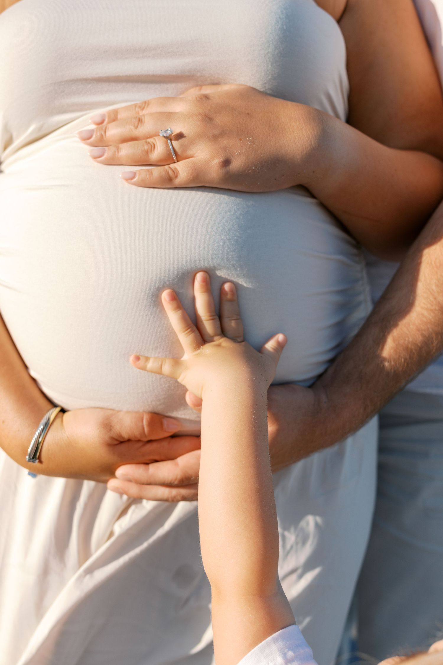 A child's hand touching a pregnant woman's belly while her partner embraces her from behind outdoors.