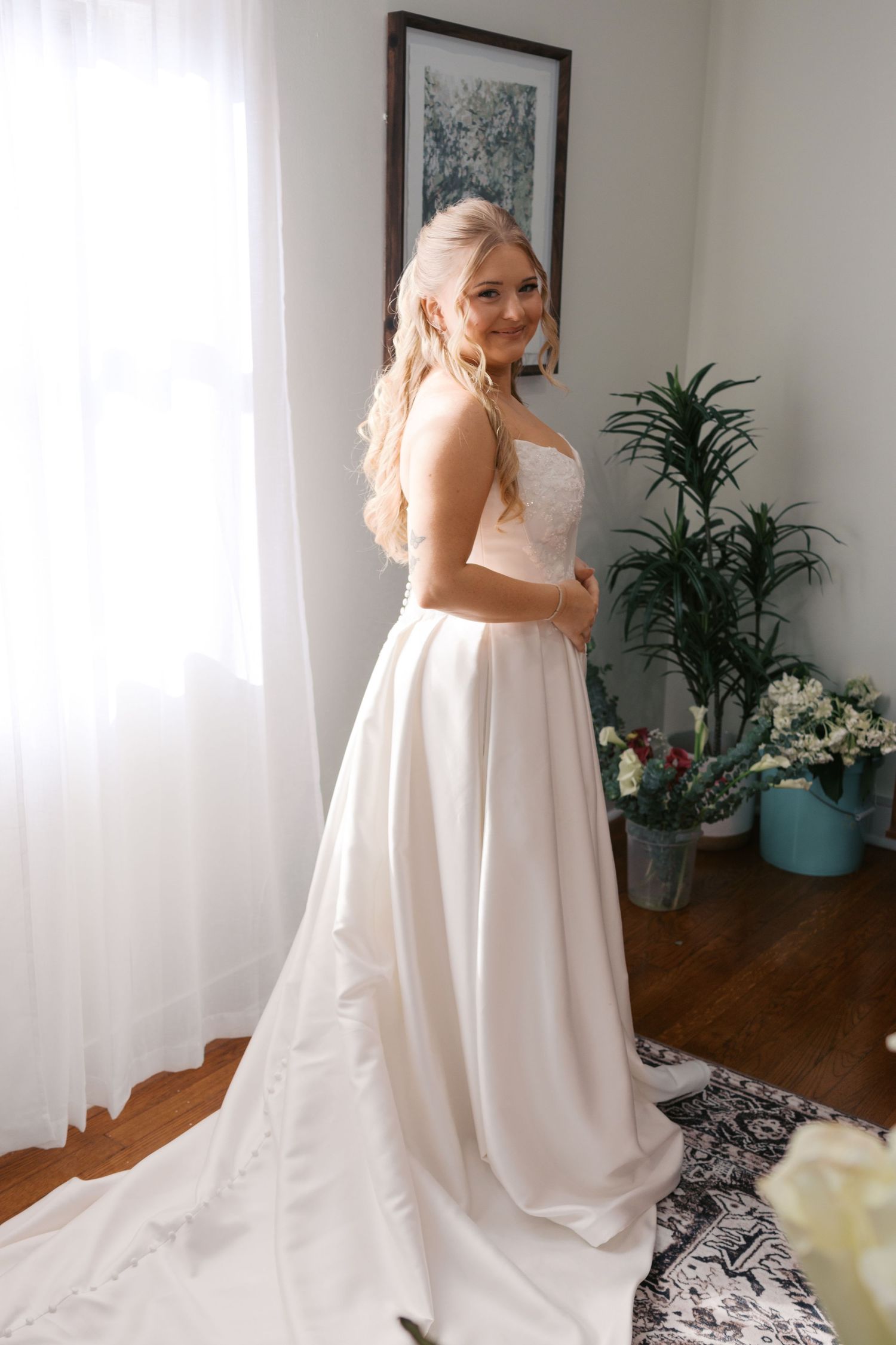 Smiling bride in elegant white satin A-line wedding gown with lace bodice posing in a bright room with plants.