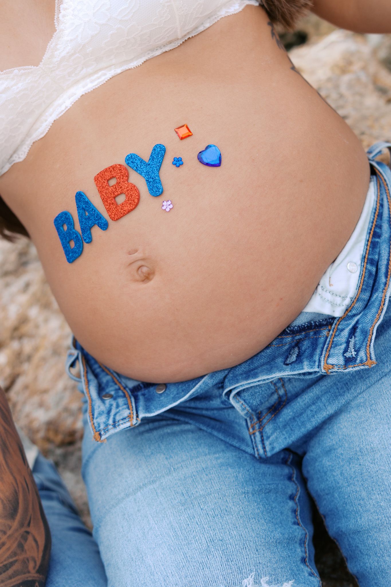 Pregnant woman's baby bump with colorful BABY letter stickers, wearing white top and distressed denim jeans.