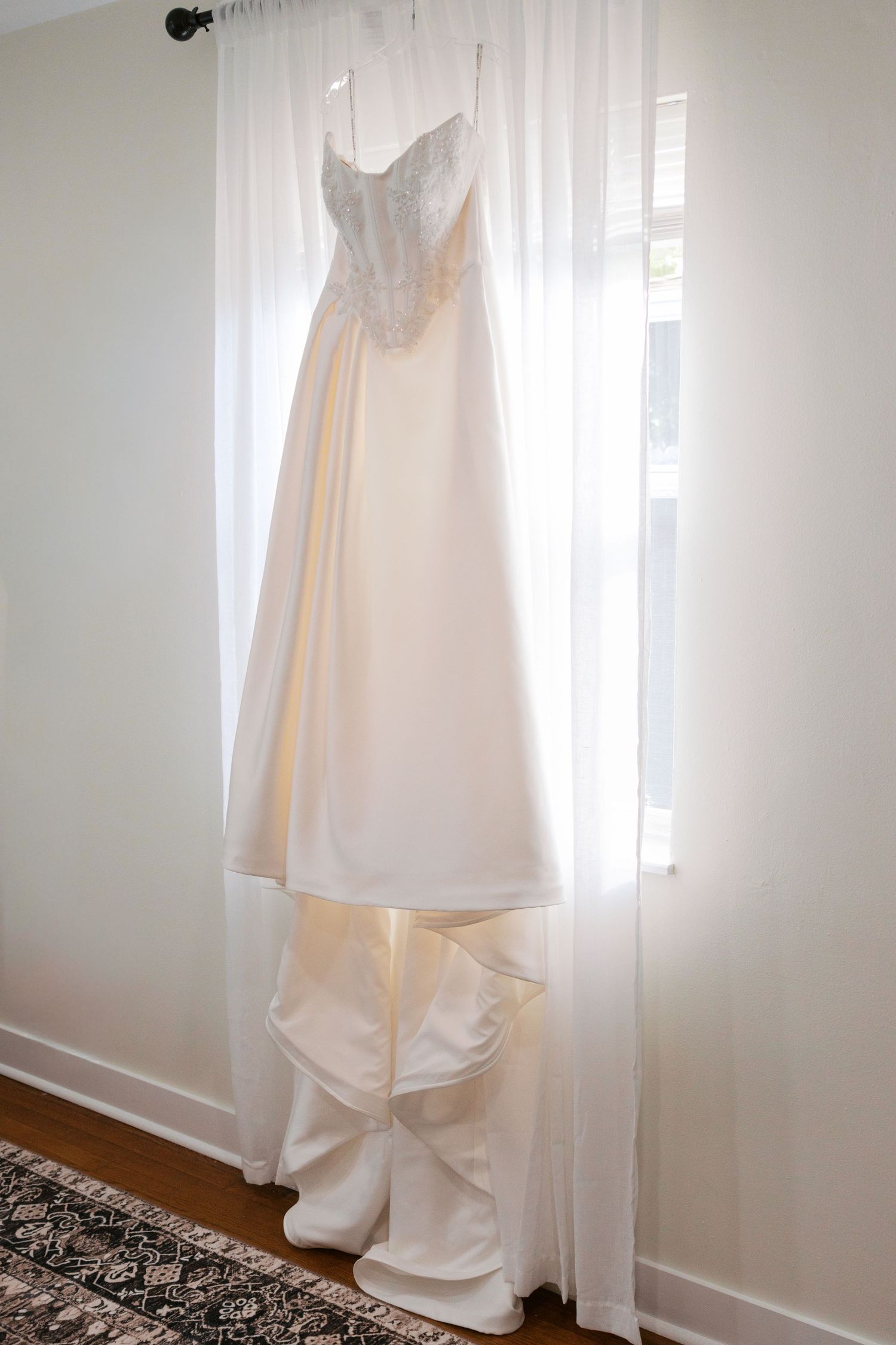 Elegant white A-line wedding dress with lace bodice hanging in front of a bright window with sheer curtains.