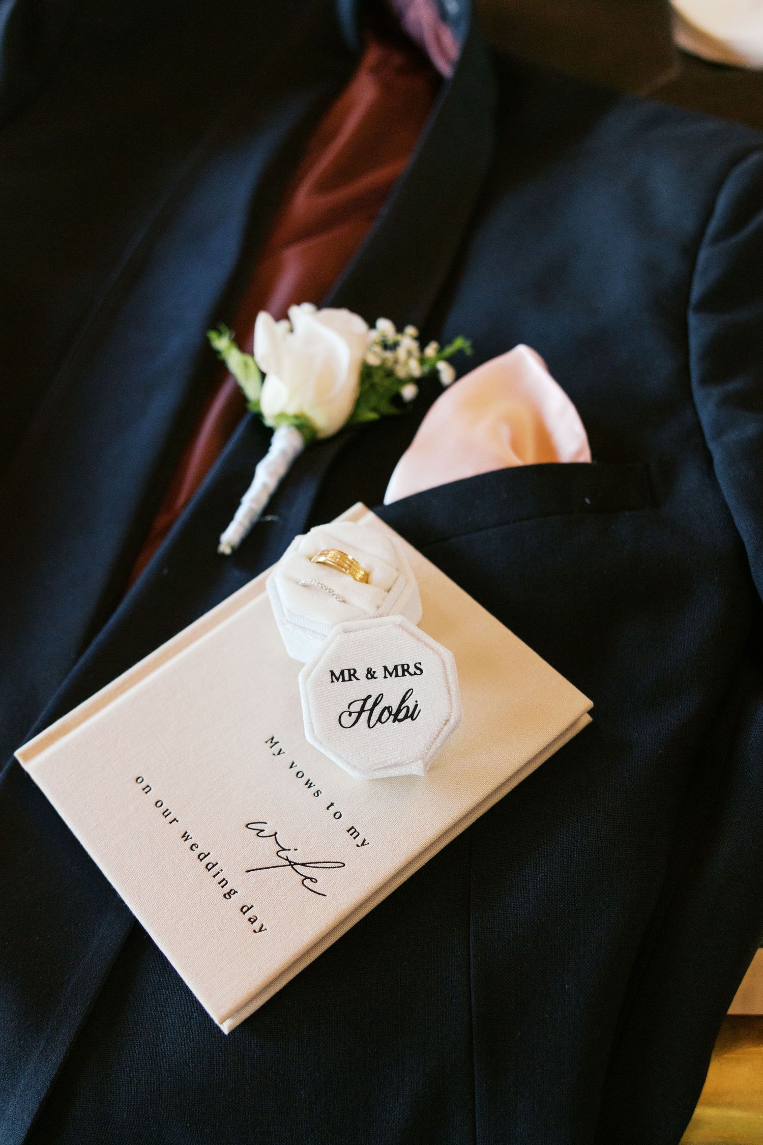 Navy suit with white rose boutonniere, pink pocket square, Mr & Mrs Hobi ring box, and wedding vow book on wedding day.