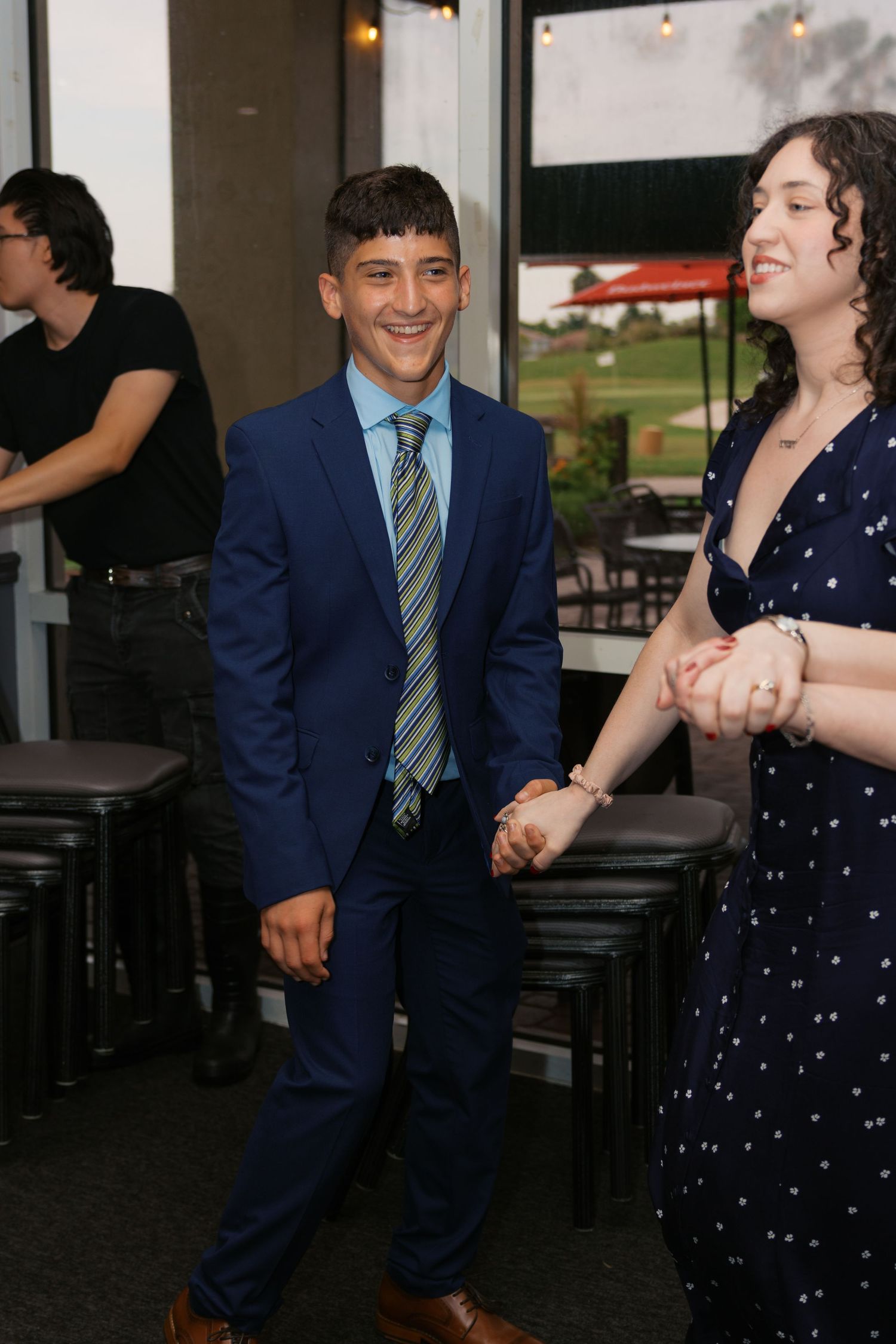 Young couple in formal attire holding hands and smiling at an indoor venue, man in blue suit with tie.