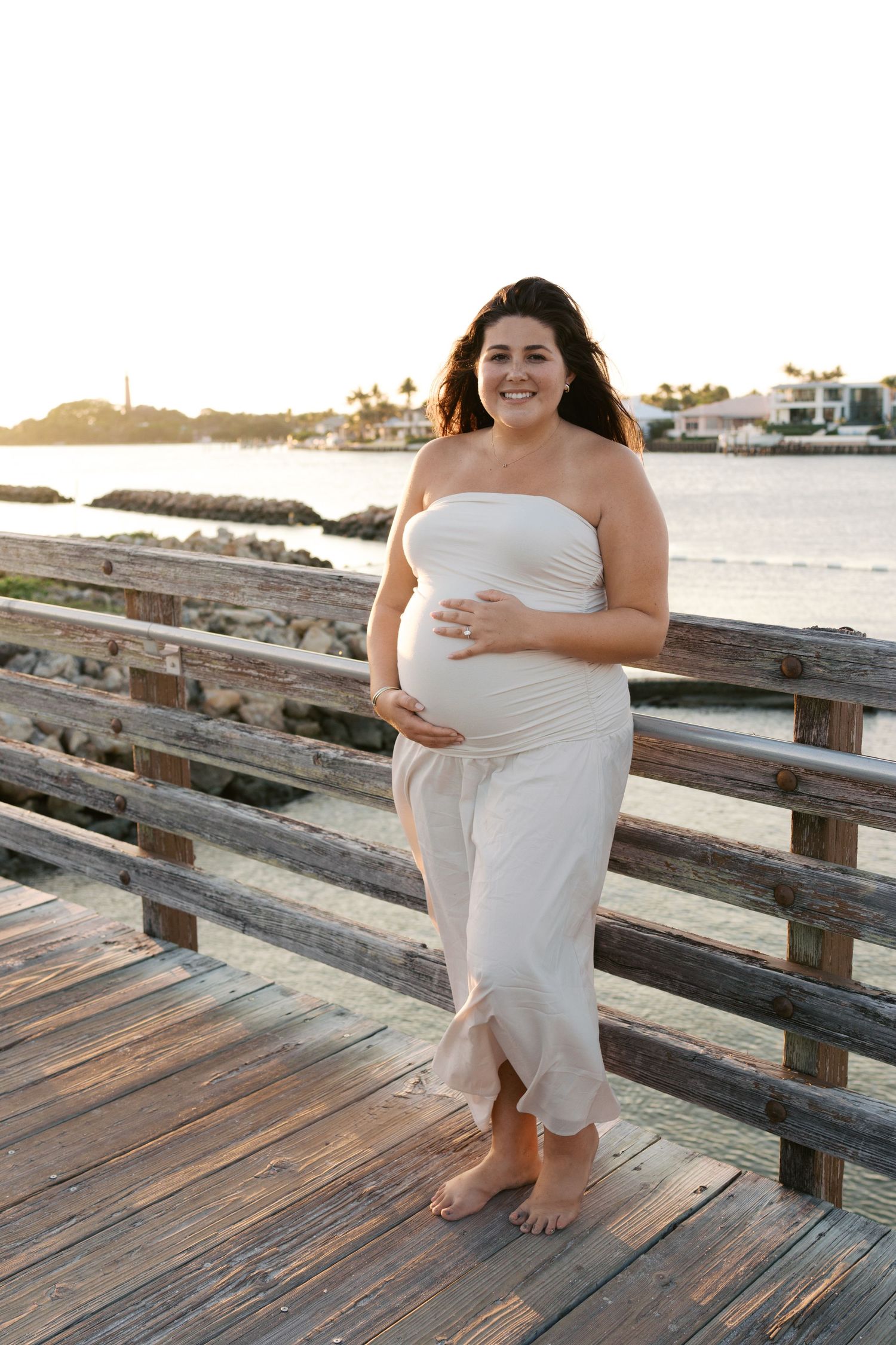 Beautiful pregnant woman in strapless white dress cradling baby bump on wooden boardwalk at golden hour sunset.