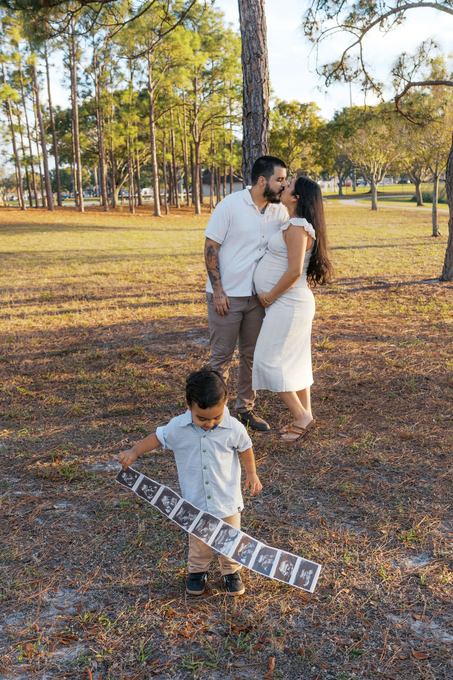 Pregnant couple kissing in park while young boy holds ultrasound photos strip during maternity photo session.