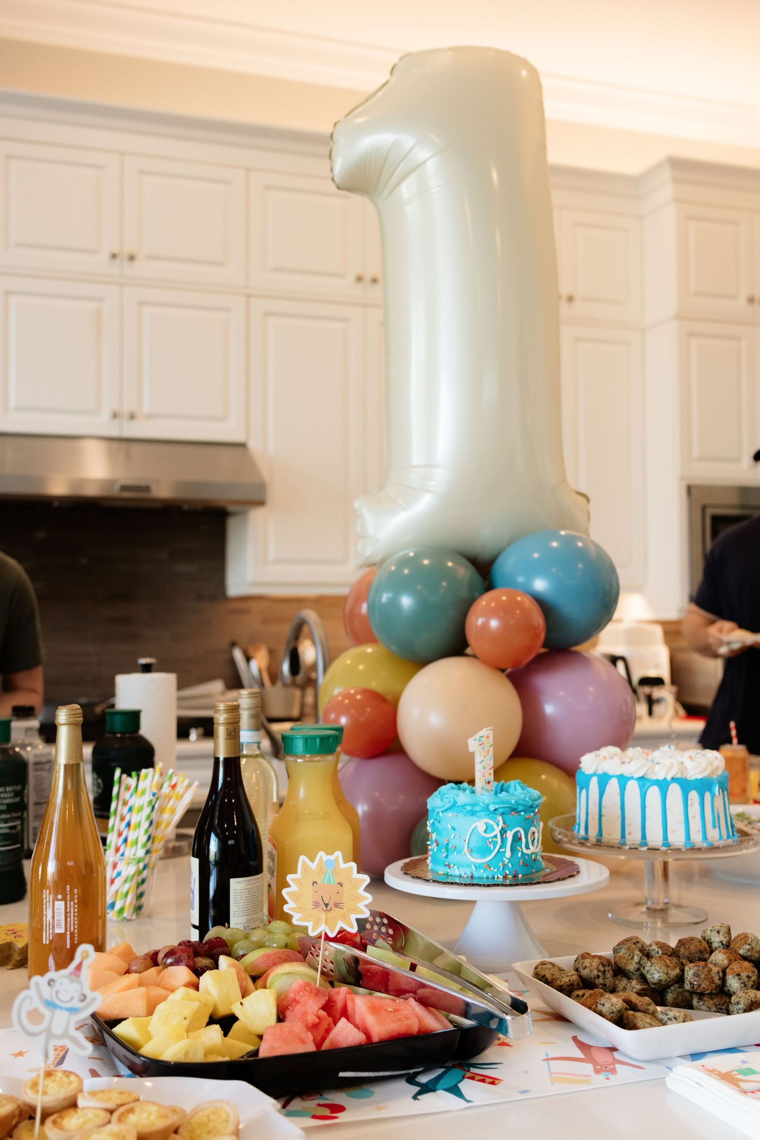 First birthday party table with number one balloon, smash cake, fruit platter, drinks and colorful balloon display.
