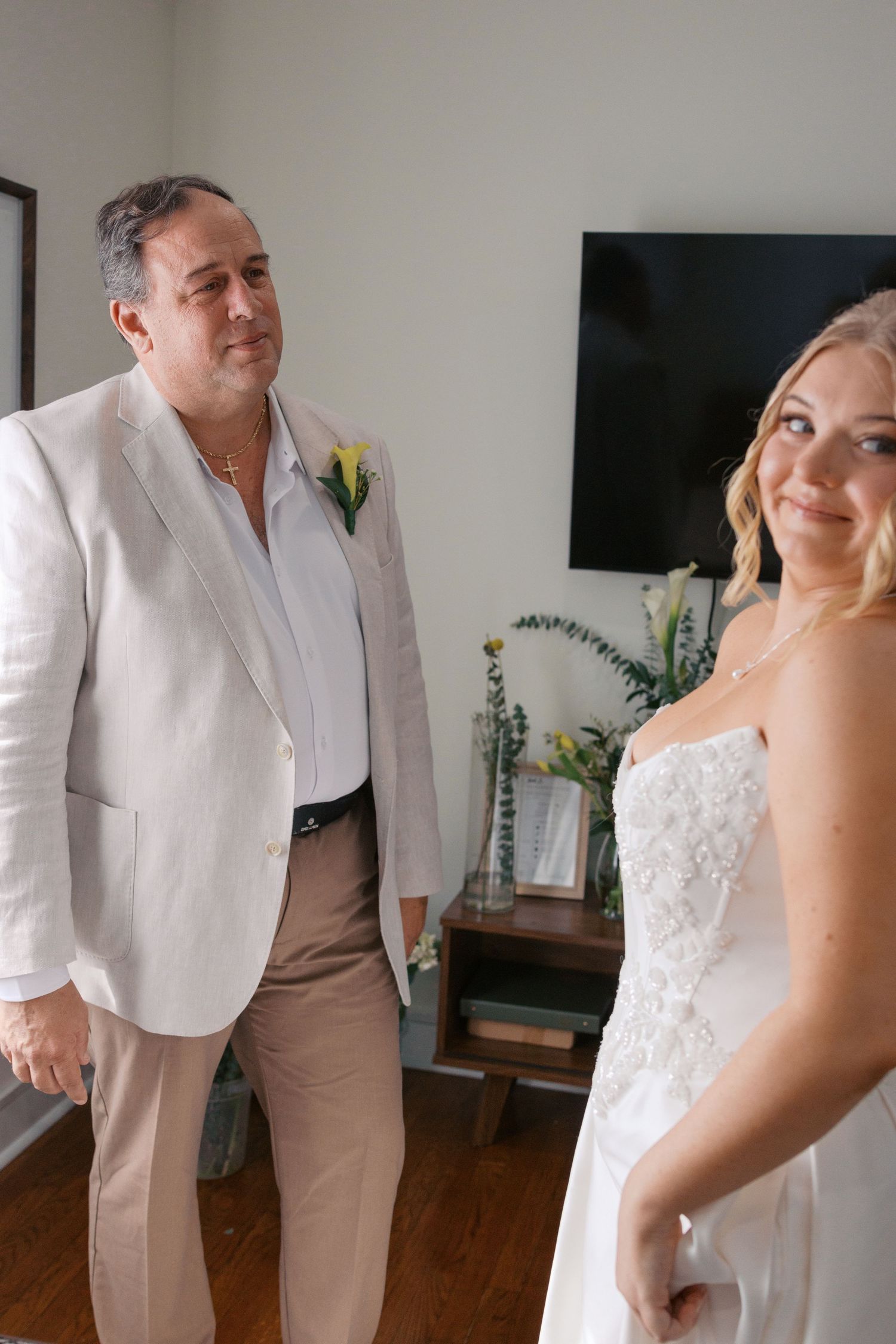 A bride in a white lace gown smiles as a man in a white suit with a yellow boutonniere looks on during wedding preparations.