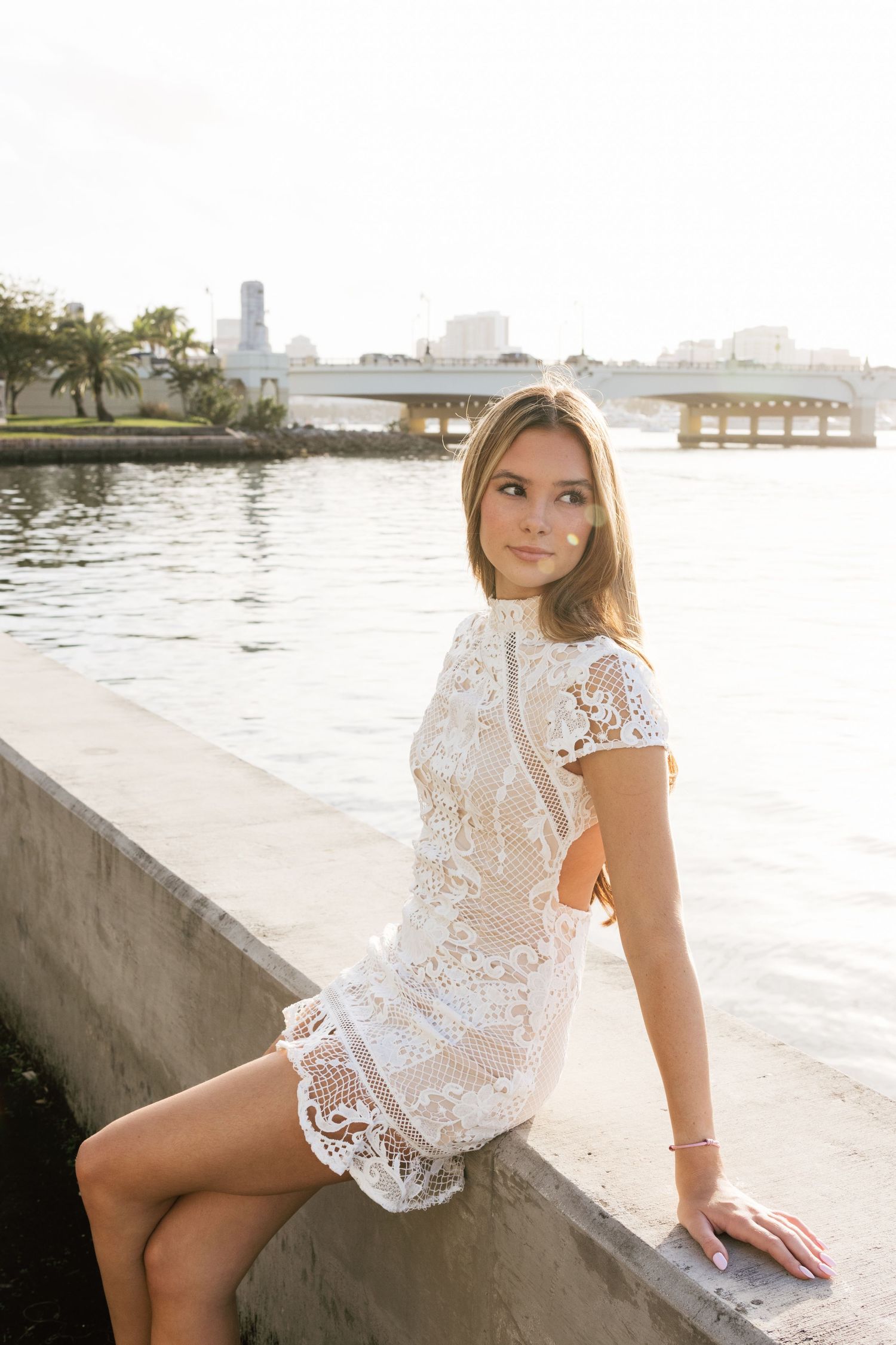 A woman in white lace poses on concrete steps near the water during golden hour.