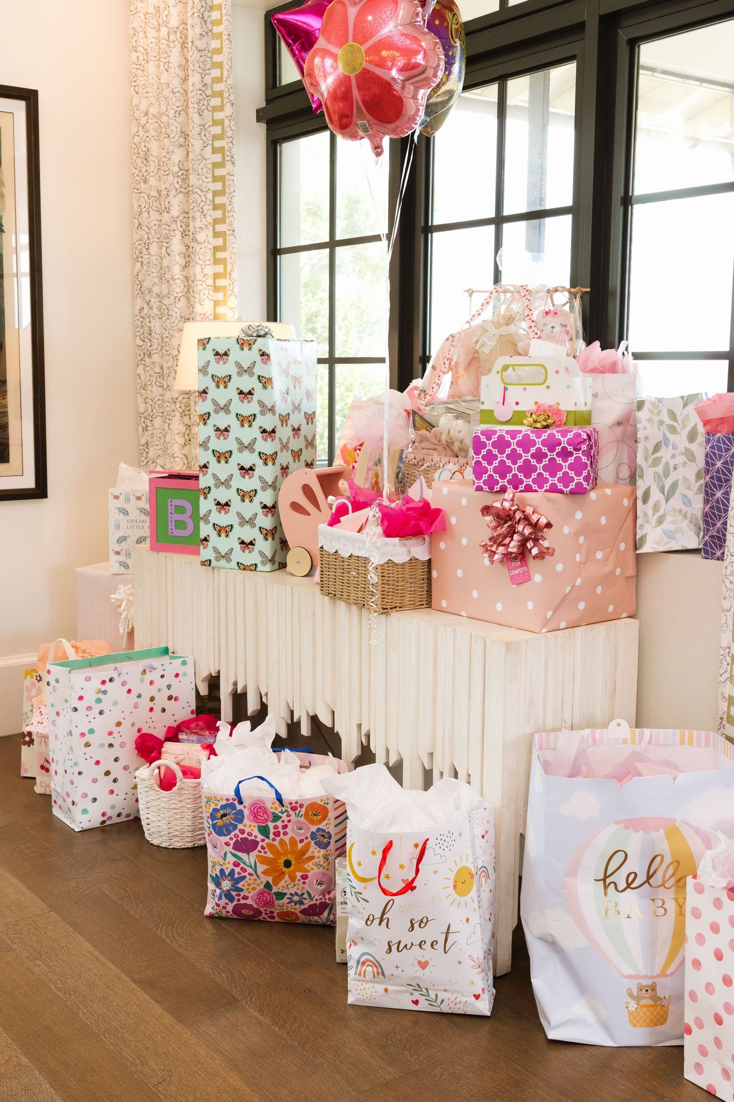 Colorful baby shower gifts and gift bags with pink tissue paper and a floral balloon displayed on a white table.