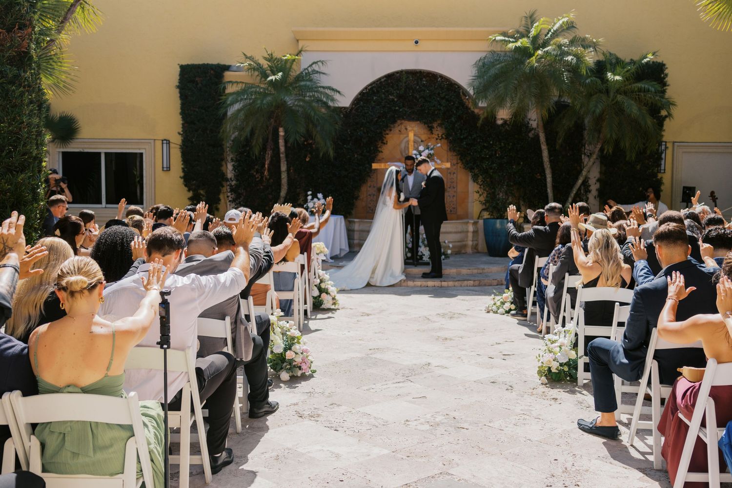 Outdoor wedding ceremony with bride and groom at altar, guests raising hands, palm trees, and stone arch backdrop.