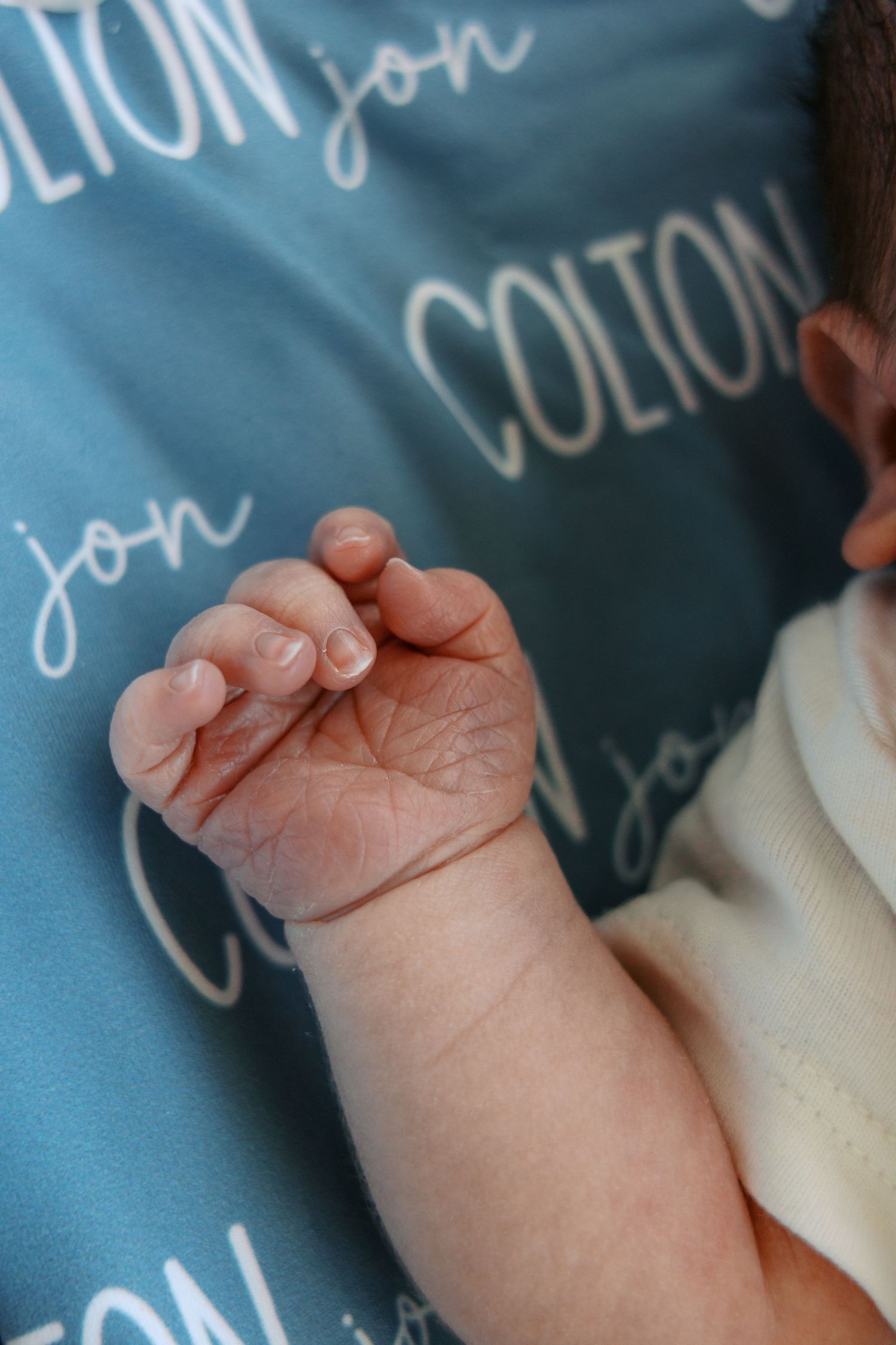 Close-up of a newborn baby's tiny hand resting on a teal personalized blanket with the name Colton Jon.