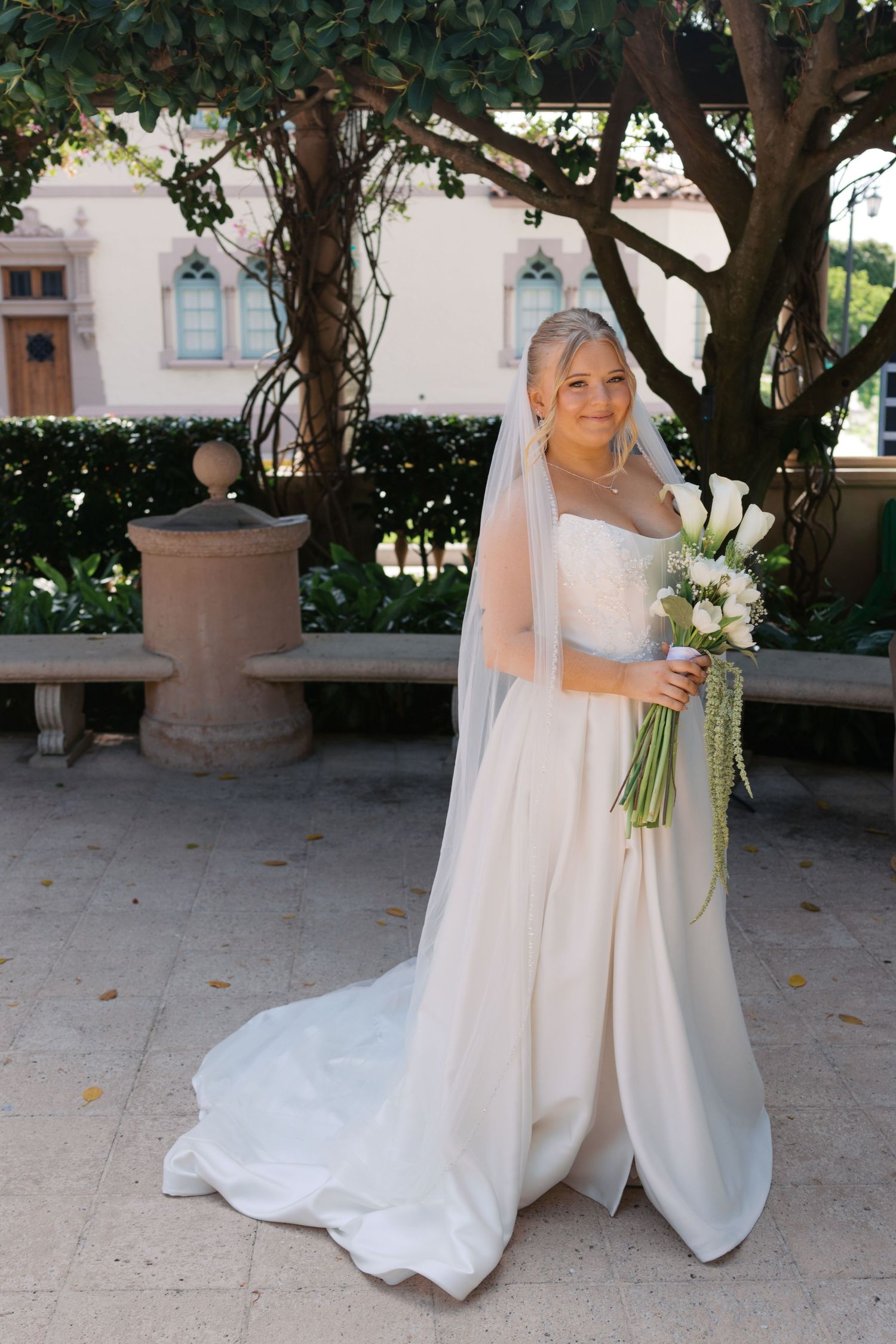 Bride in strapless white gown with veil holds white calla lily bouquet outdoors under a tree.