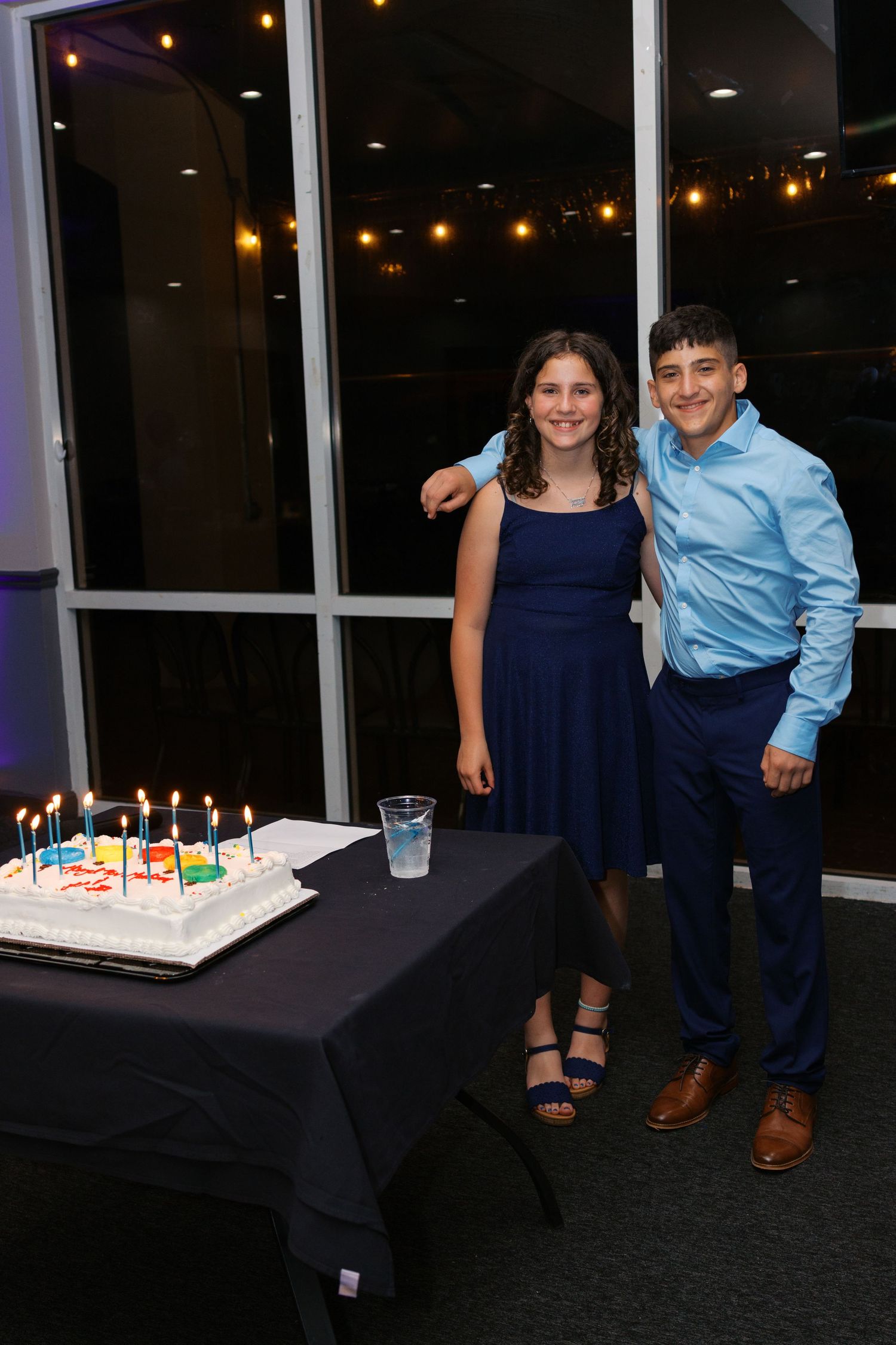 Two teens in formal attire pose beside a birthday cake with lit candles at an evening celebration.