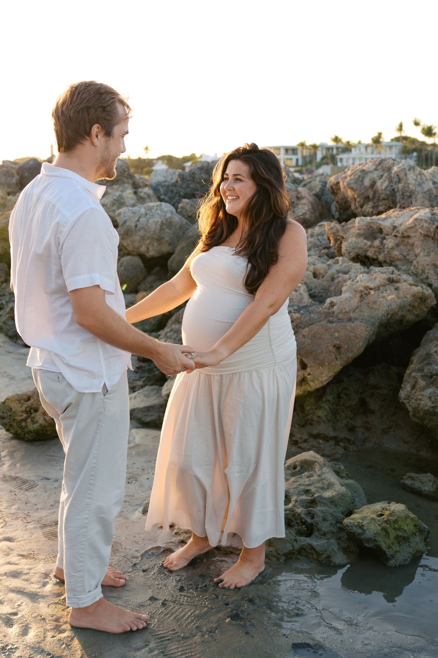 Pregnant couple holding hands on rocky beach at sunset, woman in flowing white maternity dress.