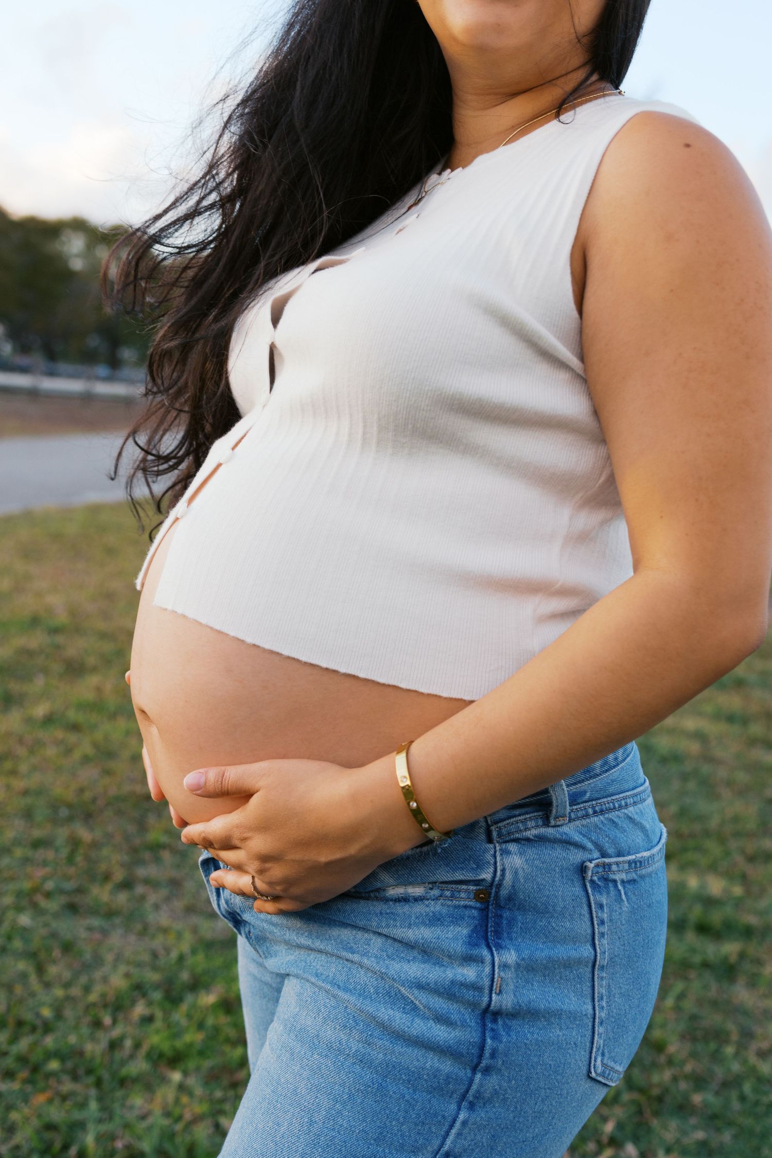 Pregnant woman in white crop top and jeans cradling baby bump outdoors at golden hour.