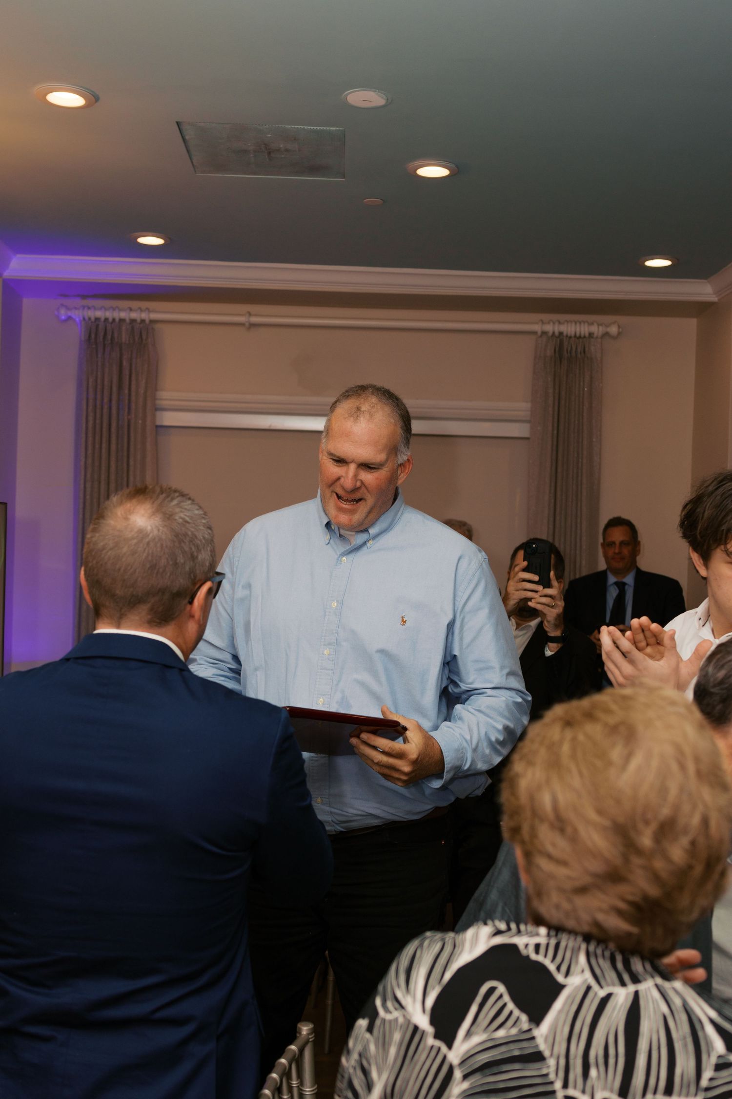 Man in blue shirt receives award plaque at indoor ceremony while audience applauds in elegant event venue.