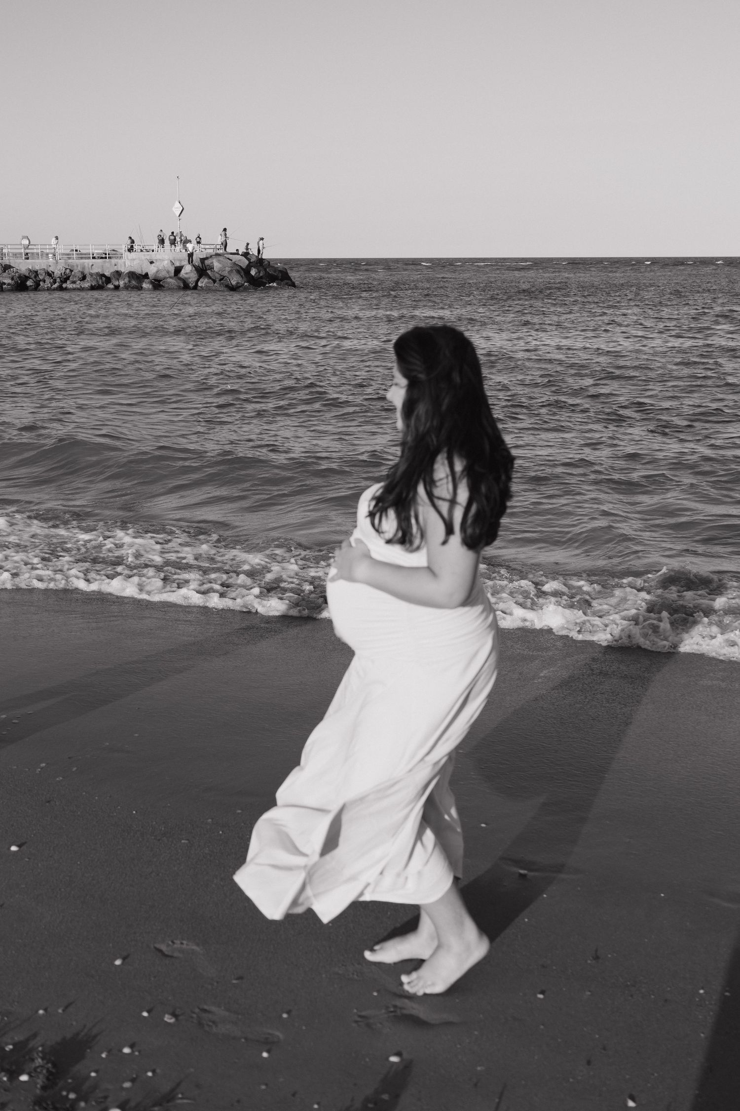 Black and white photo of a woman in a flowing white dress walking barefoot along a sandy beach near ocean waves.